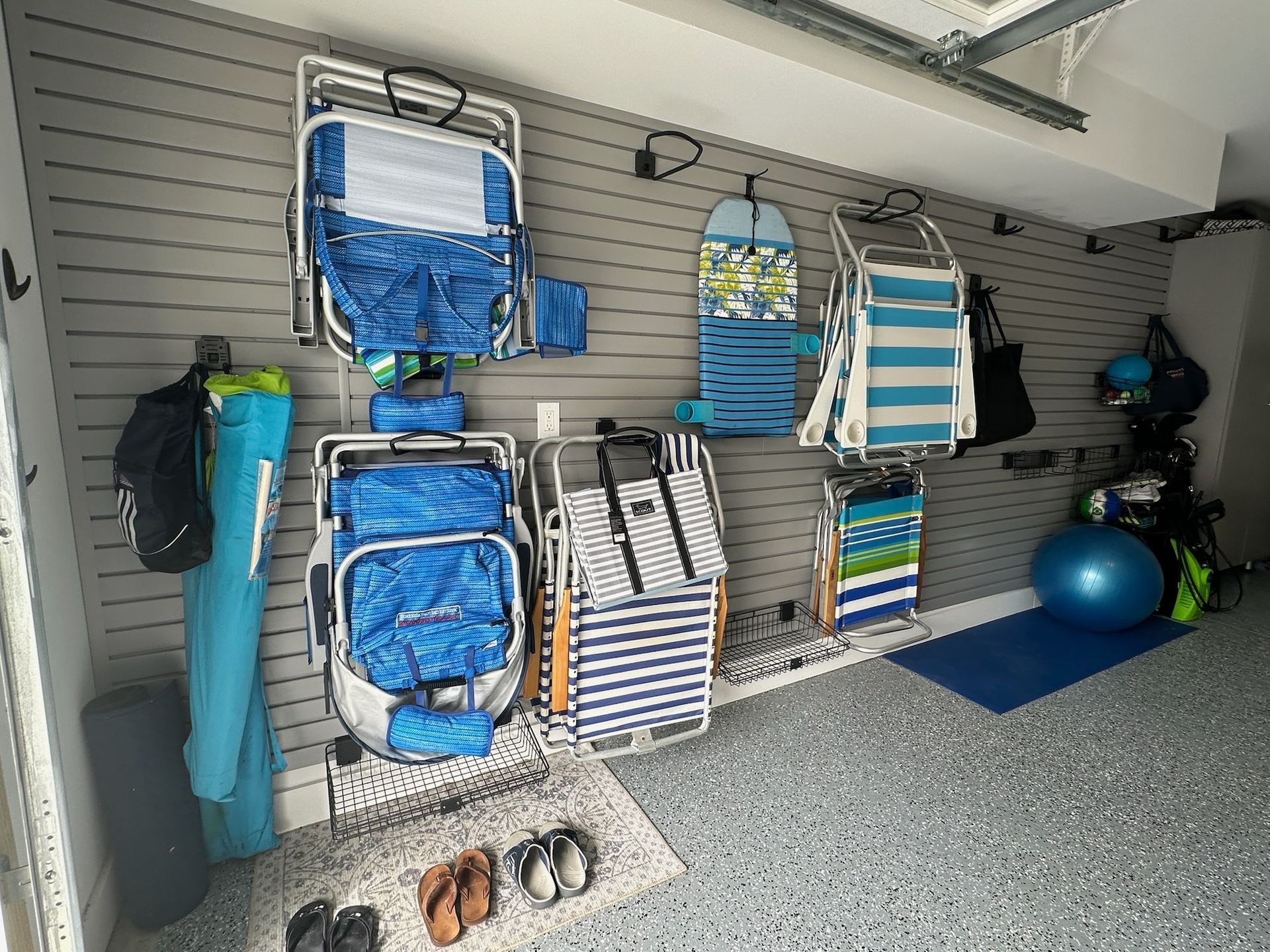 Beach chairs and gear organized on a slatted wall in a garage, with sandals on the floor and a blue exercise ball nearby.