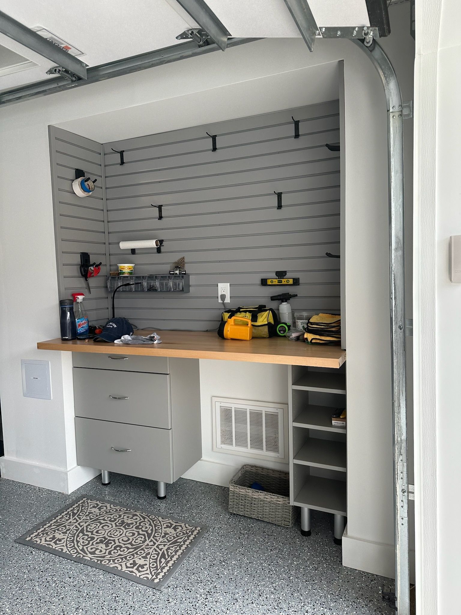 A grey slat-wall workbench with a wooden top, two-drawer cabinet, and open shelving in a garage with speckled flooring.
