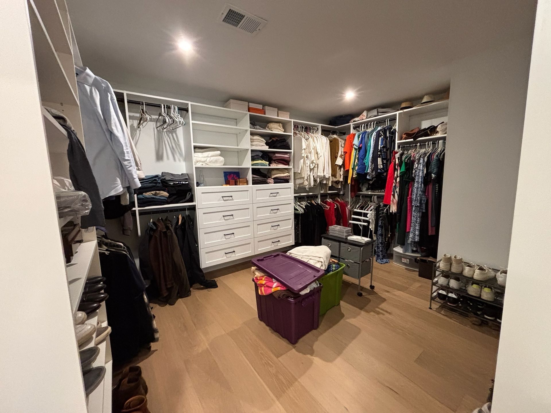 A large, organized walk-in closet with hanging clothes, shelves, white drawers, and storage bins on a light wood floor.