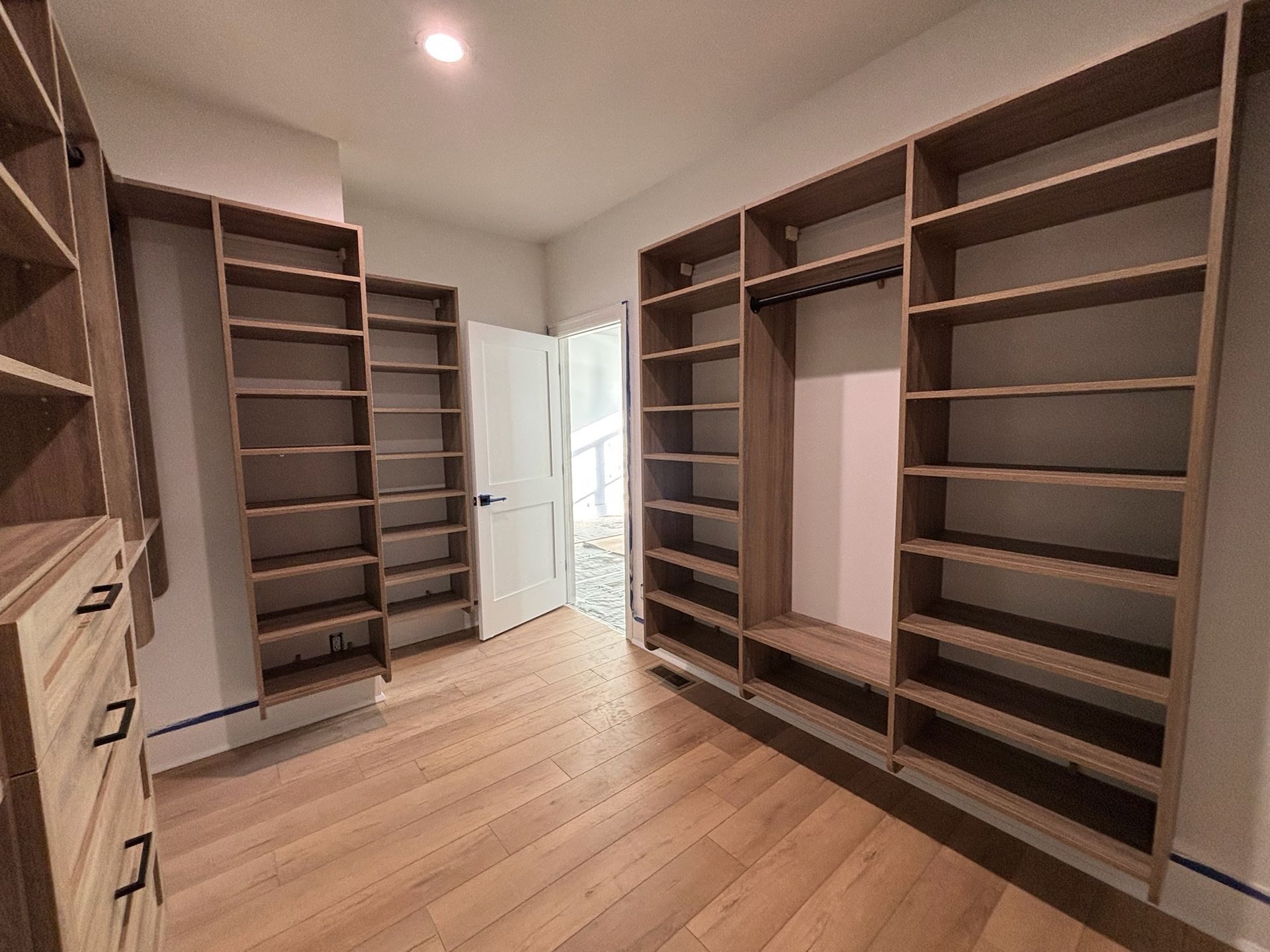A walk-in closet featuring light wood flooring, matching wooden shelving units, and a white door leading to another room.