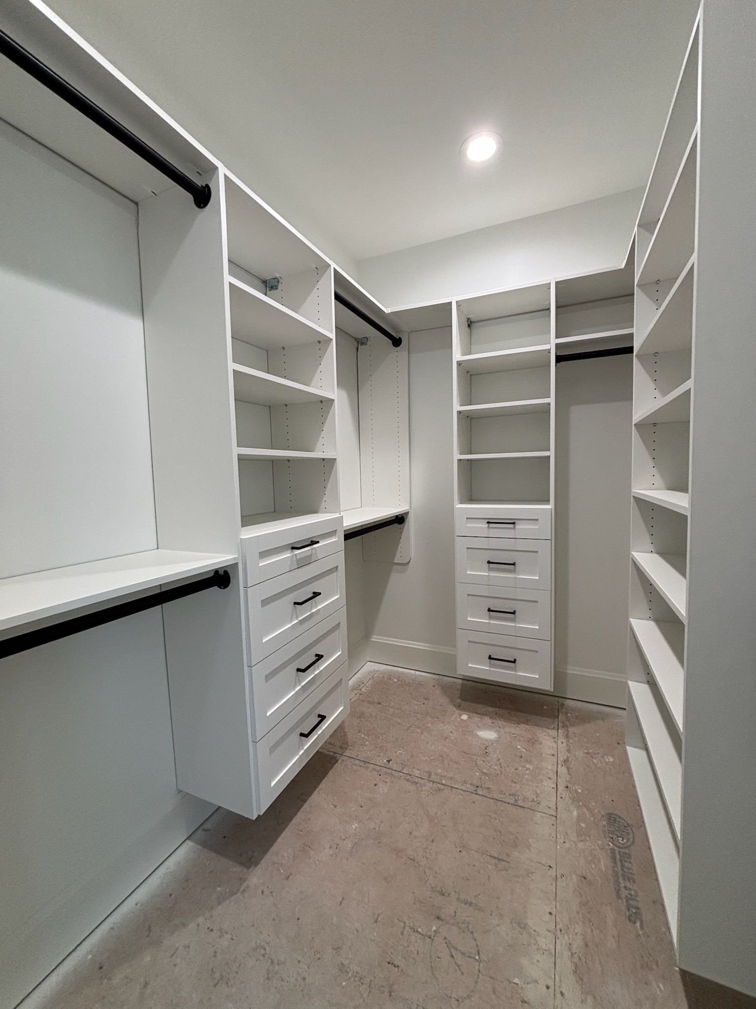 A walk-in closet featuring white wooden storage units, shelving, drawers, and black hanging rods on a neutral-toned floor.