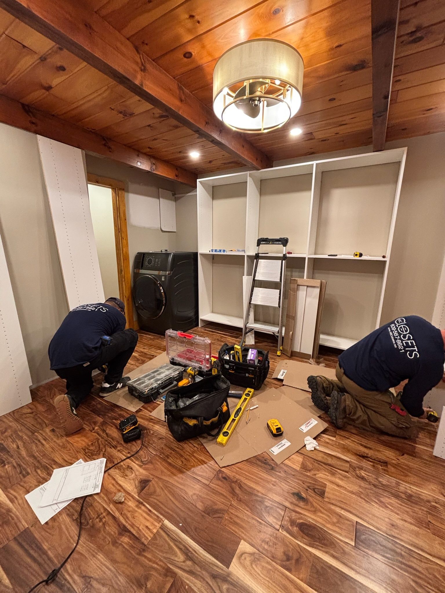 Two people in dark shirts work on installing white custom cabinetry in a room with wood-paneled ceilings and plank flooring.