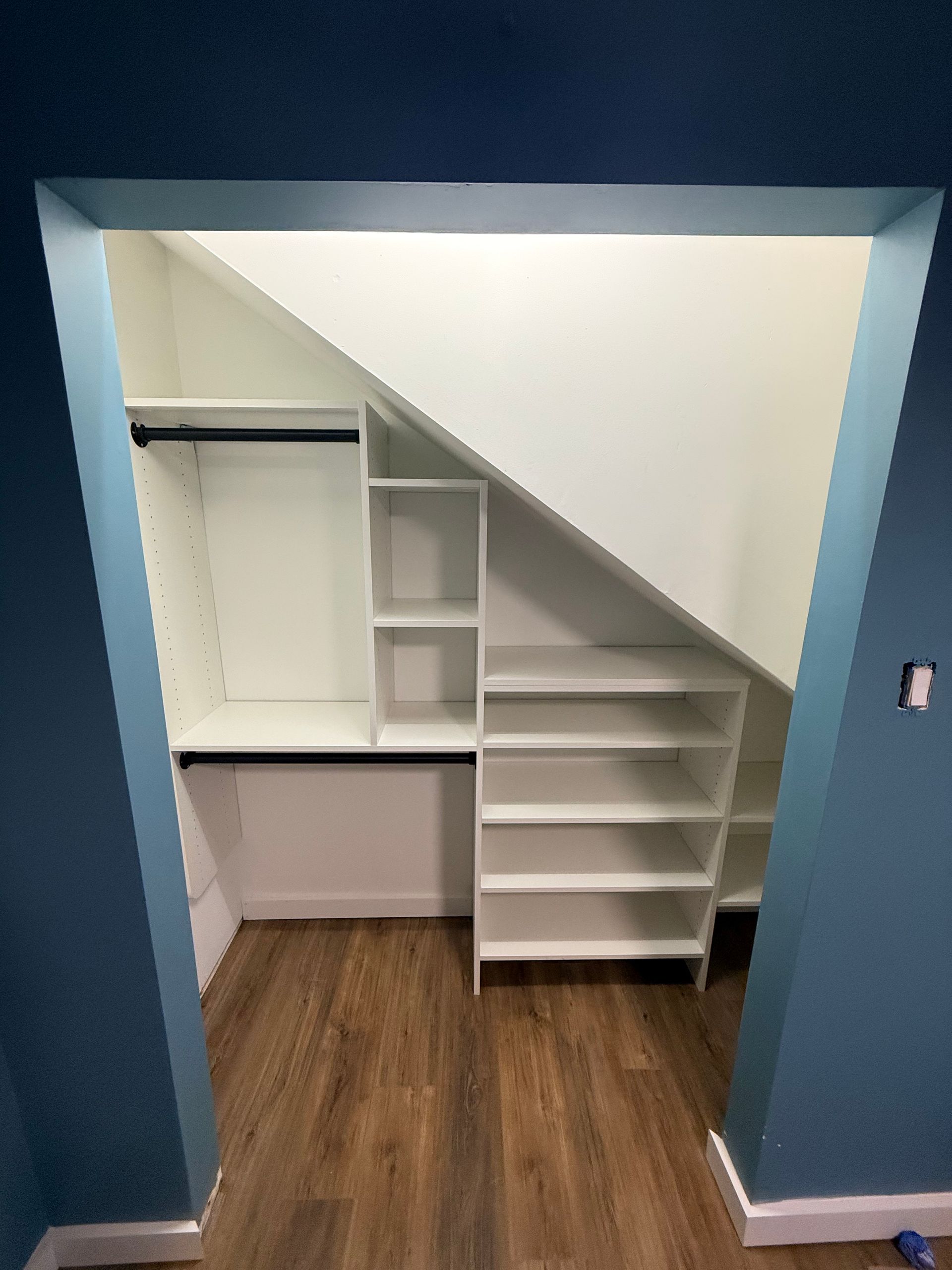 Built-in white shelving and hanging rods inside an under-stair closet with dark blue trim and wood-look flooring.