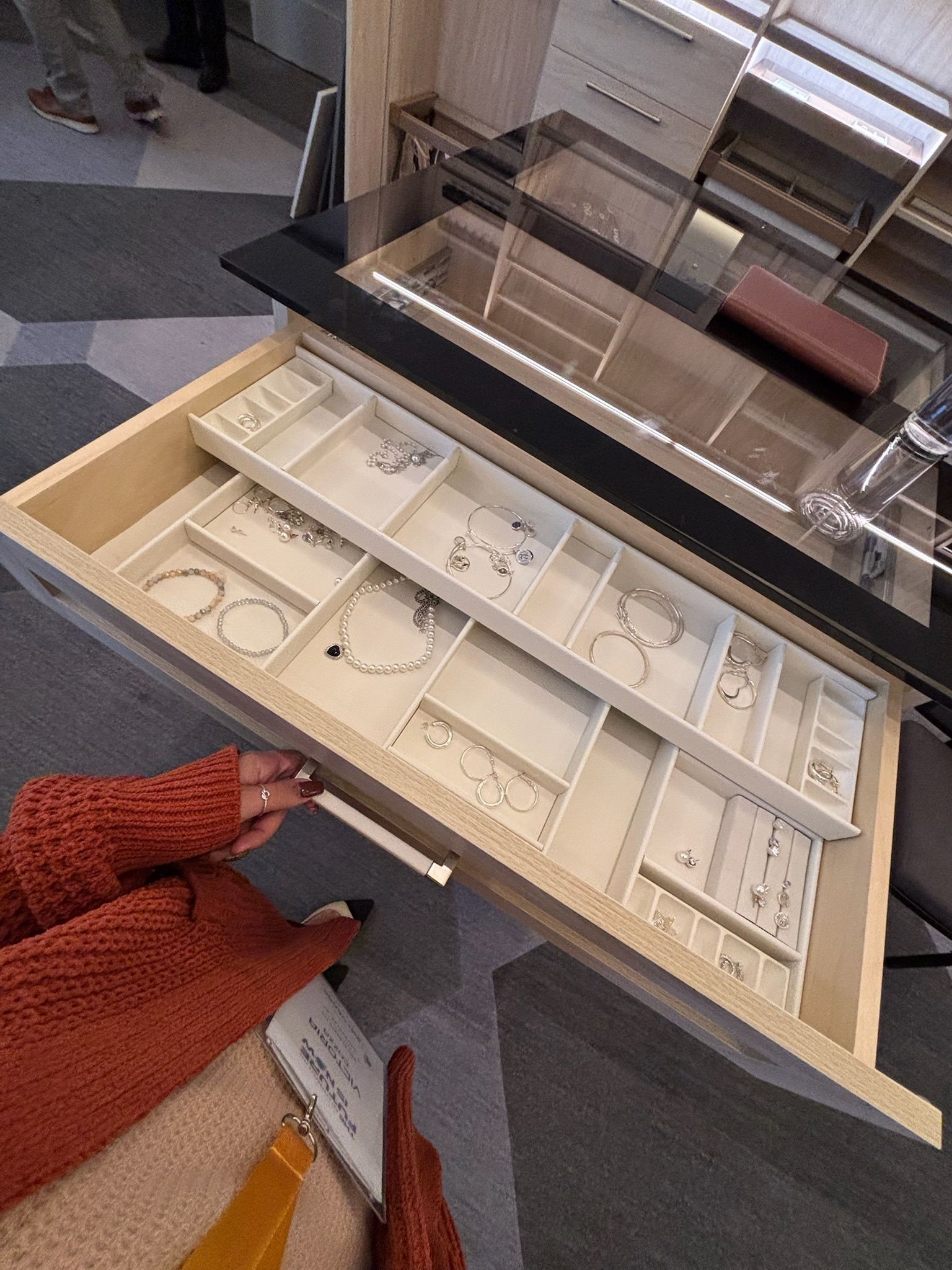 A person in a rust-colored sweater opens a light wood jewelry drawer with organized compartments and hanging jewelry.