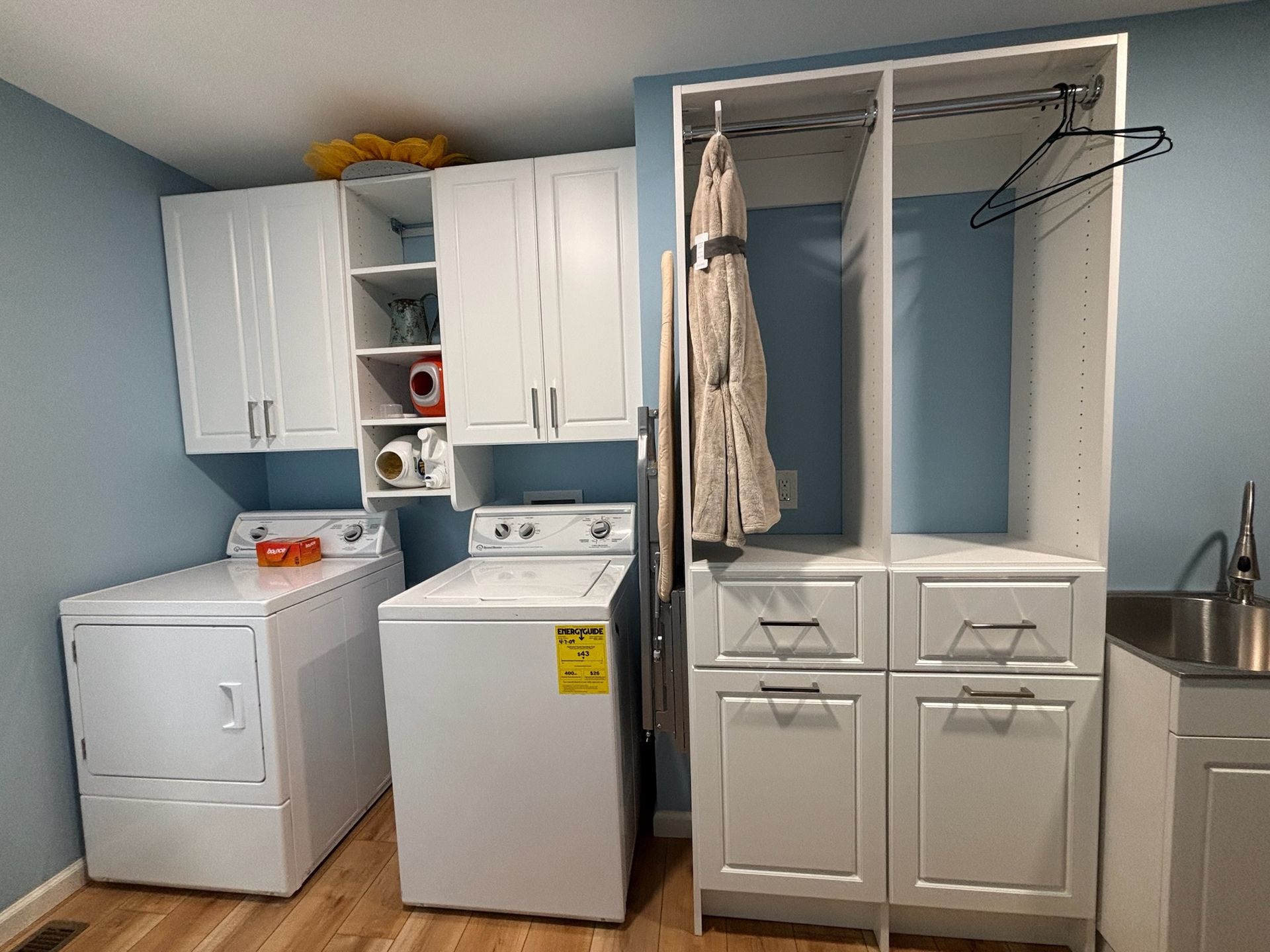 A laundry room with a white washer and dryer, upper cabinets, a tall storage unit with hanging clothes, and a sink.