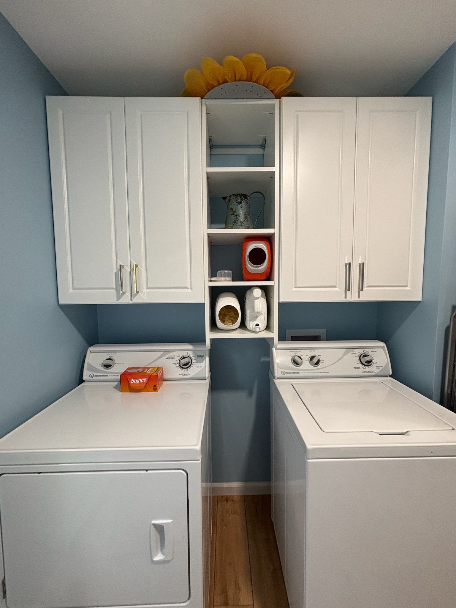 A laundry room with a white washer and dryer, center shelving unit, and white cabinets against light blue walls.