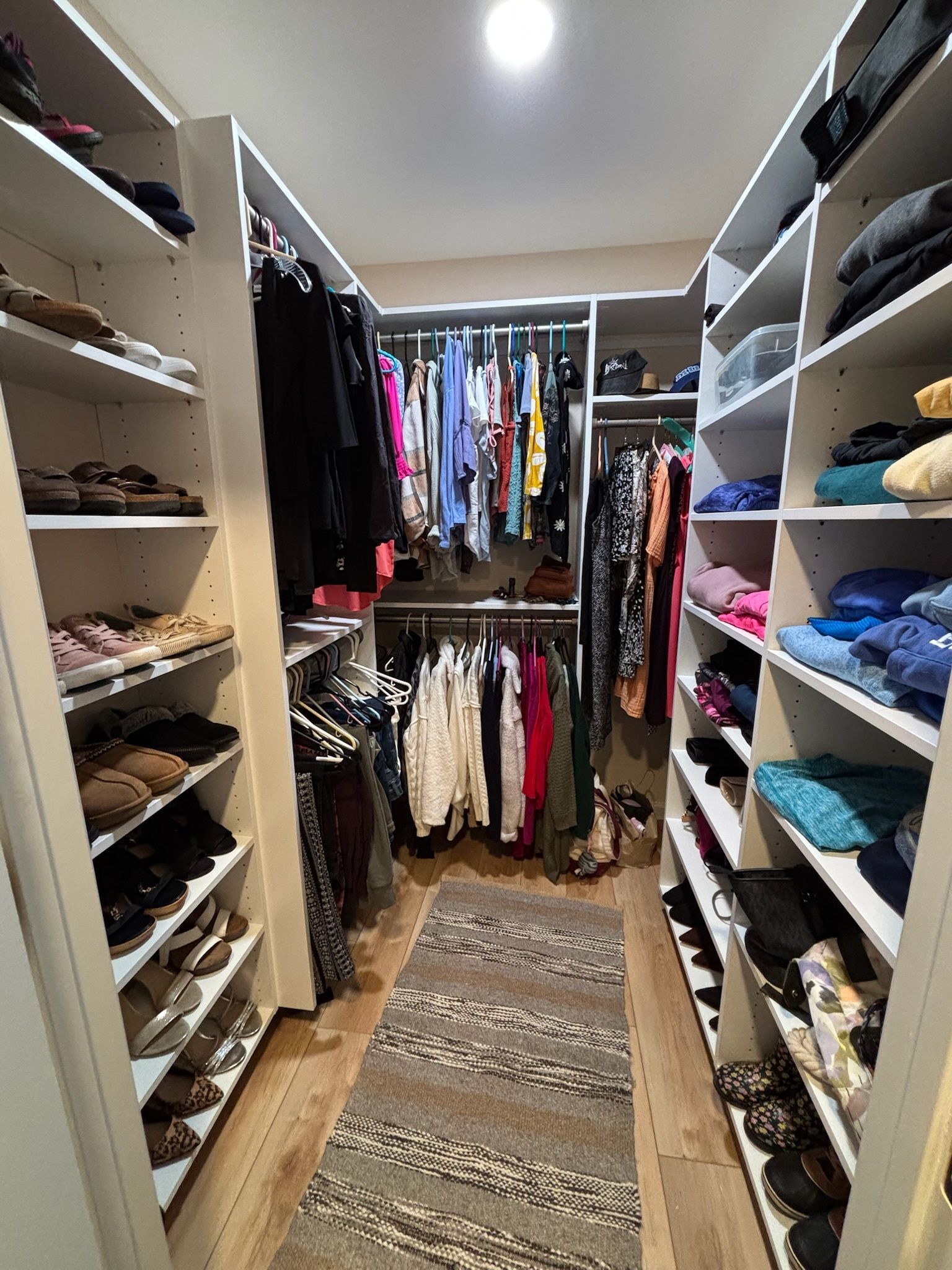 A walk-in closet with shelves full of shoes and folded clothes, and hanging garments on racks along the walls.
