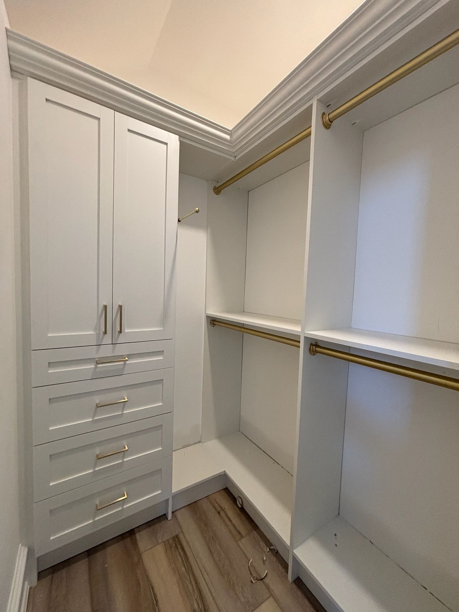 A walk-in closet with white cabinets, drawers, shelving, and gold-toned hanging rods on a wood-look floor.