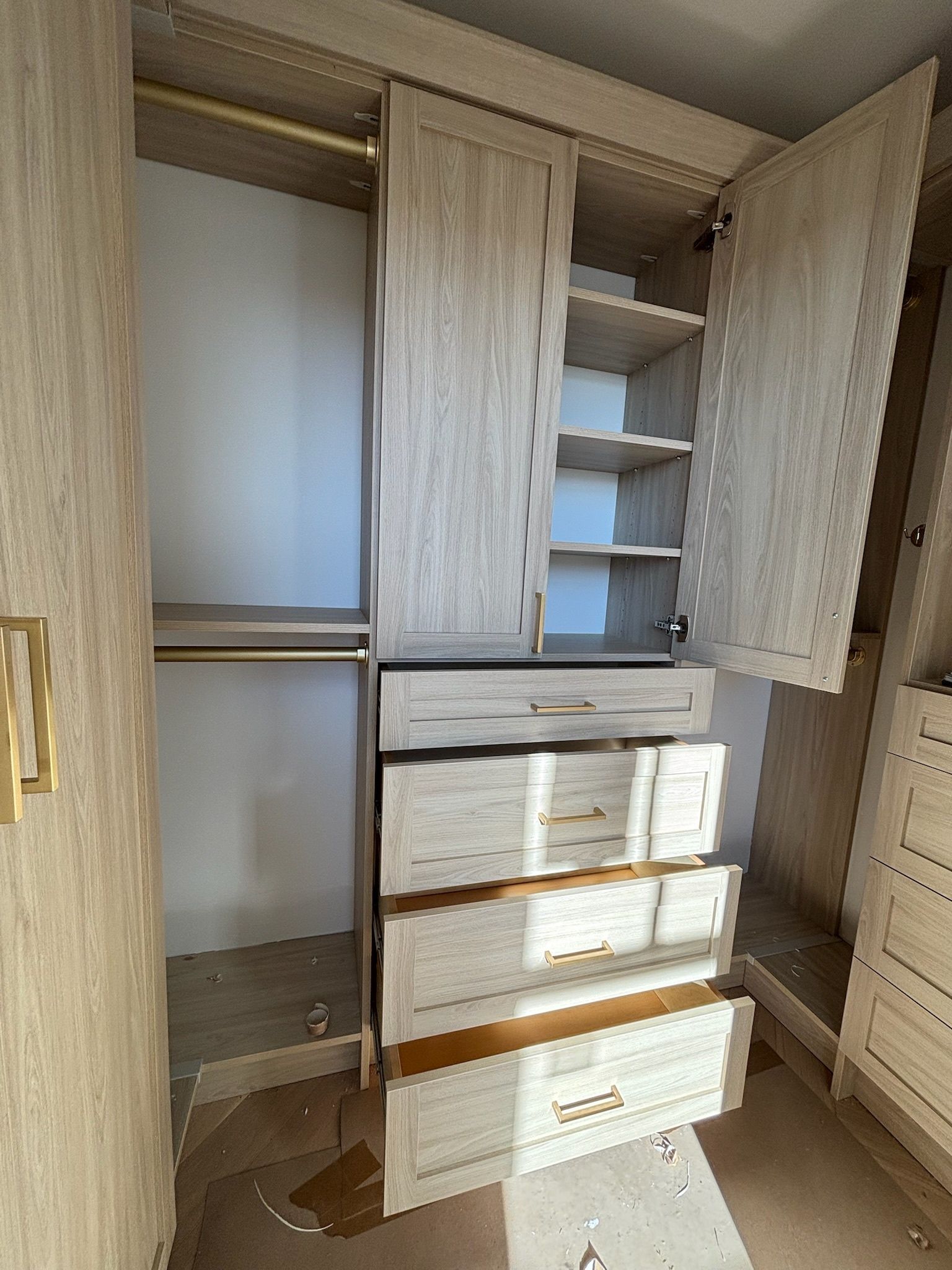 A light wood-toned wardrobe unit with an open hanging area, open shelves, and four drawers in a room under construction.