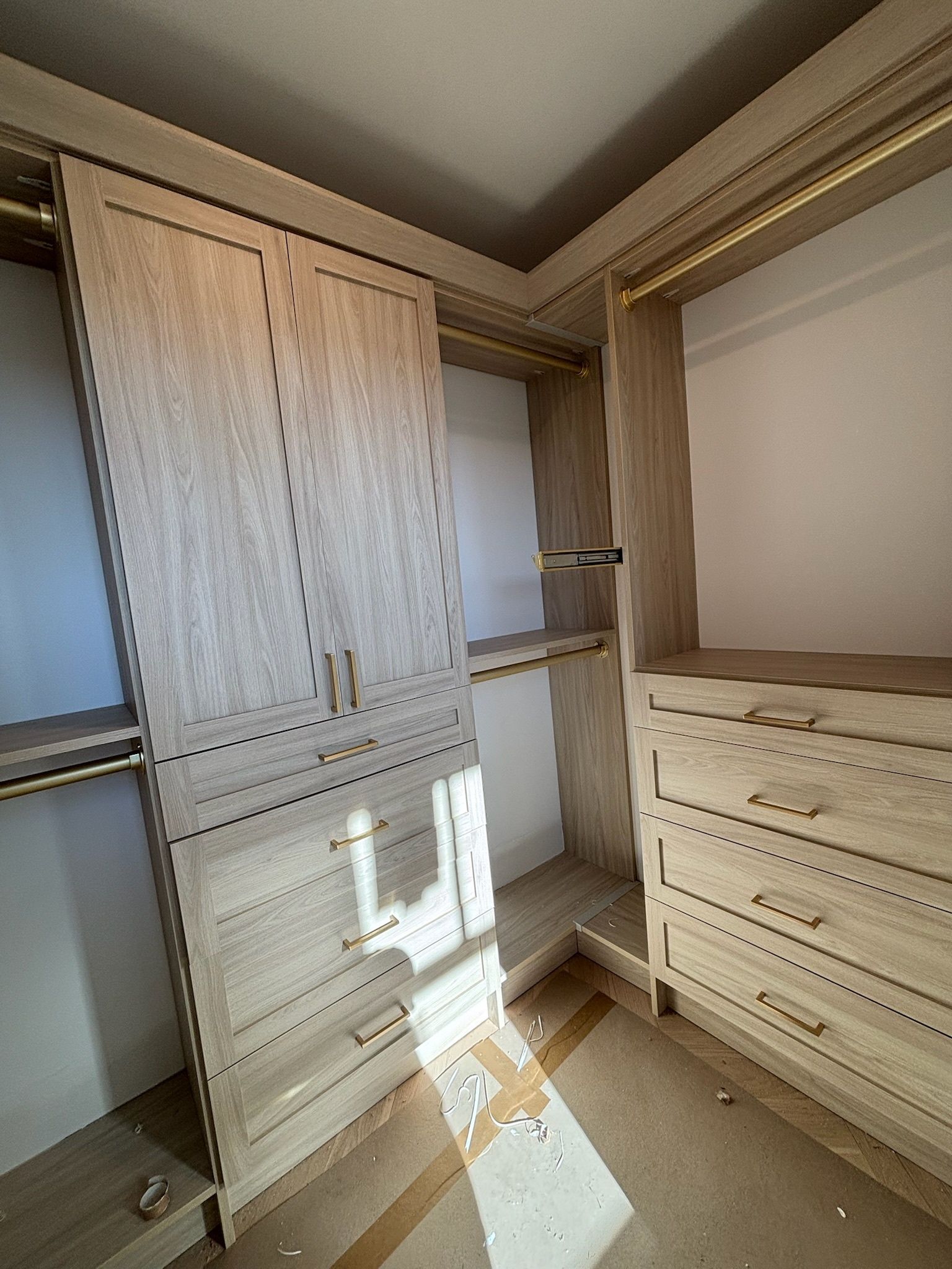 A modern walk-in closet featuring light wood cabinets, drawers, and shelving with gold hardware under bright lighting.