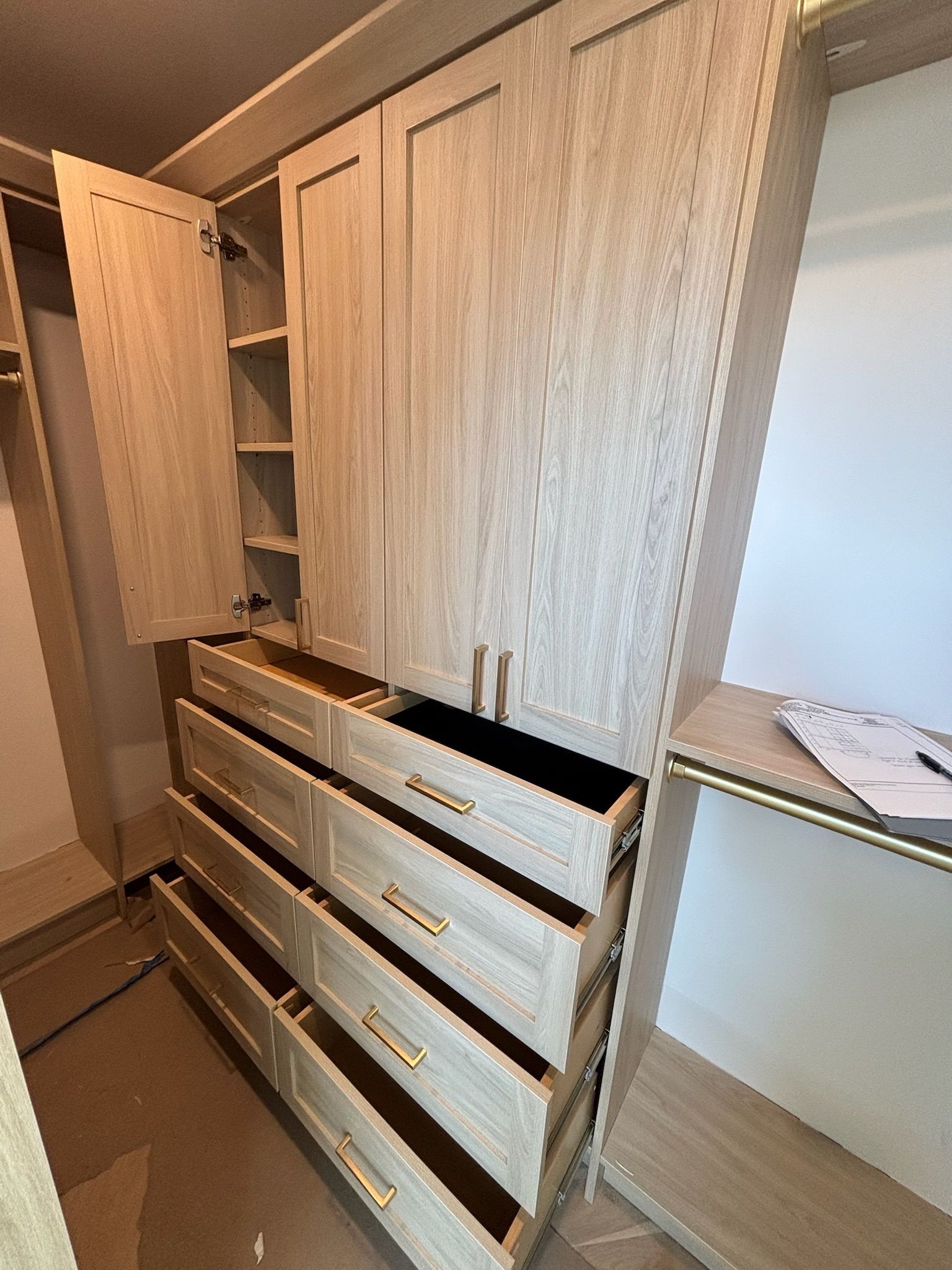 Light-colored wood closet unit with open drawers and cabinet doors, featuring gold hardware in a home setting.