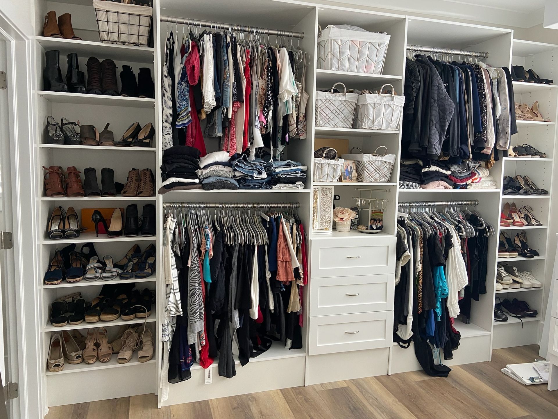 A large, white, organized walk-in closet filled with hanging clothes, stacked shelves, drawers, and many pairs of shoes.