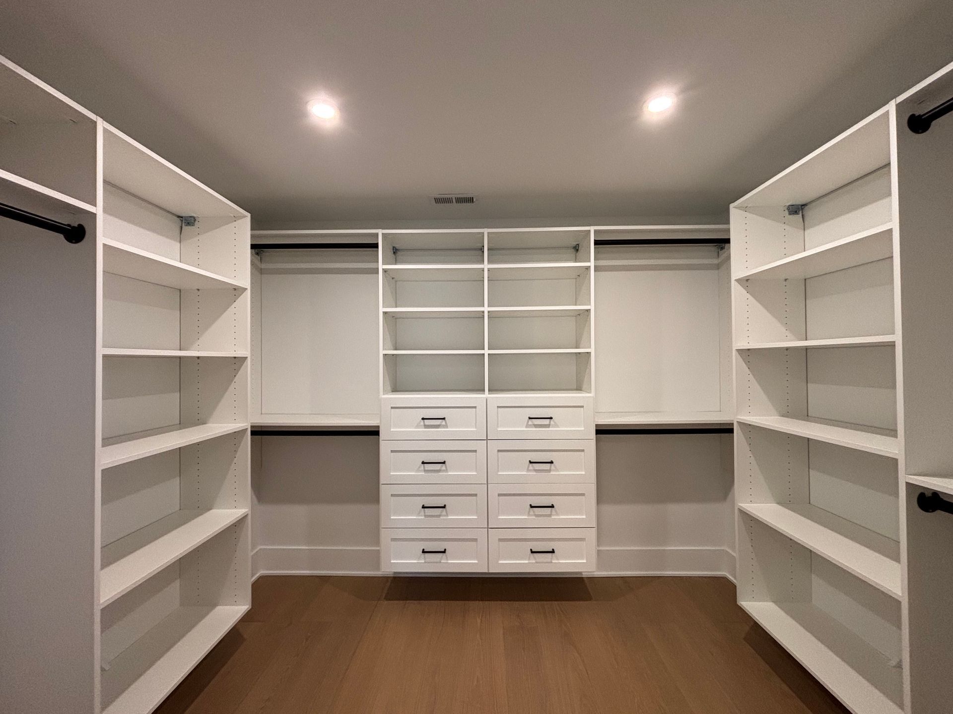 Walk-in closet with white shelving, hanging rods, and a central cabinet unit with drawers against a light gray wall.