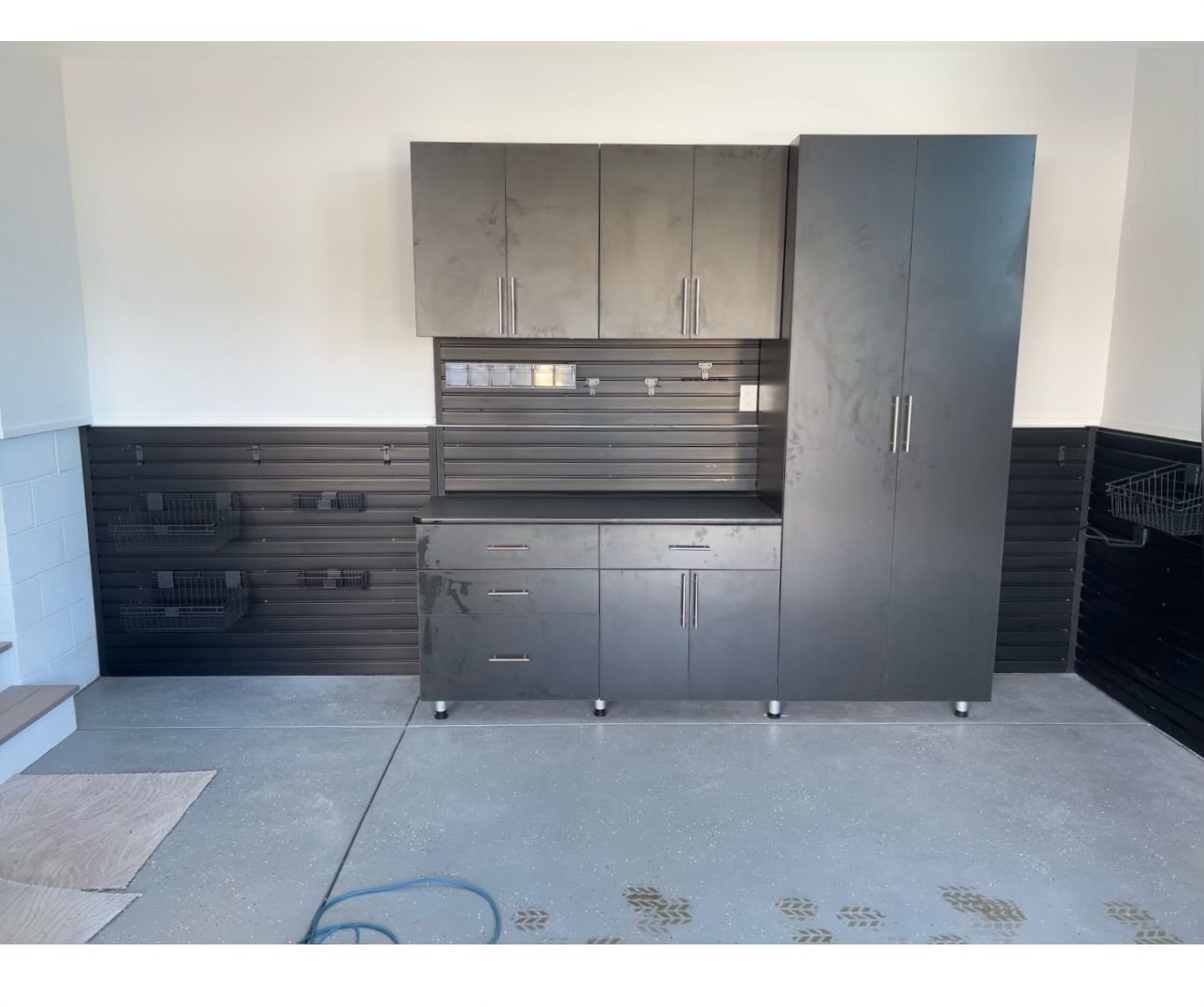 A dark gray garage storage system with wall-mounted cabinets, a countertop, drawers, and a tall locker on a concrete floor.