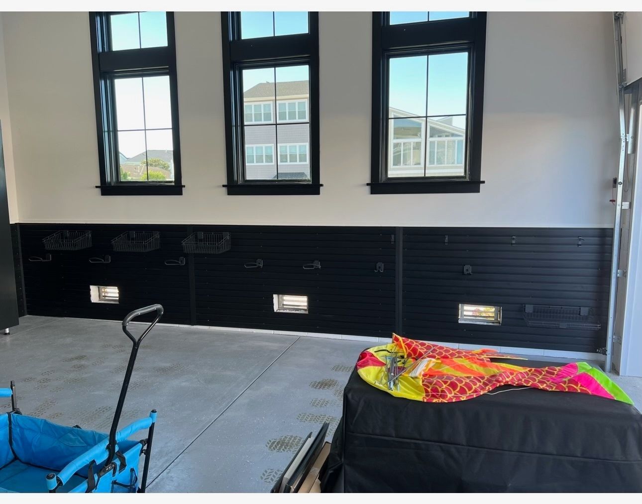 A garage interior features three windows above a black slatted storage wall, a blue utility cart, and a covered item.