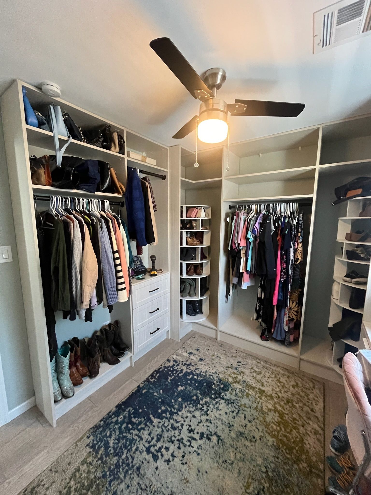 A walk-in closet featuring built-in white shelving, organized clothing, hanging garments, and a textured blue-green rug.
