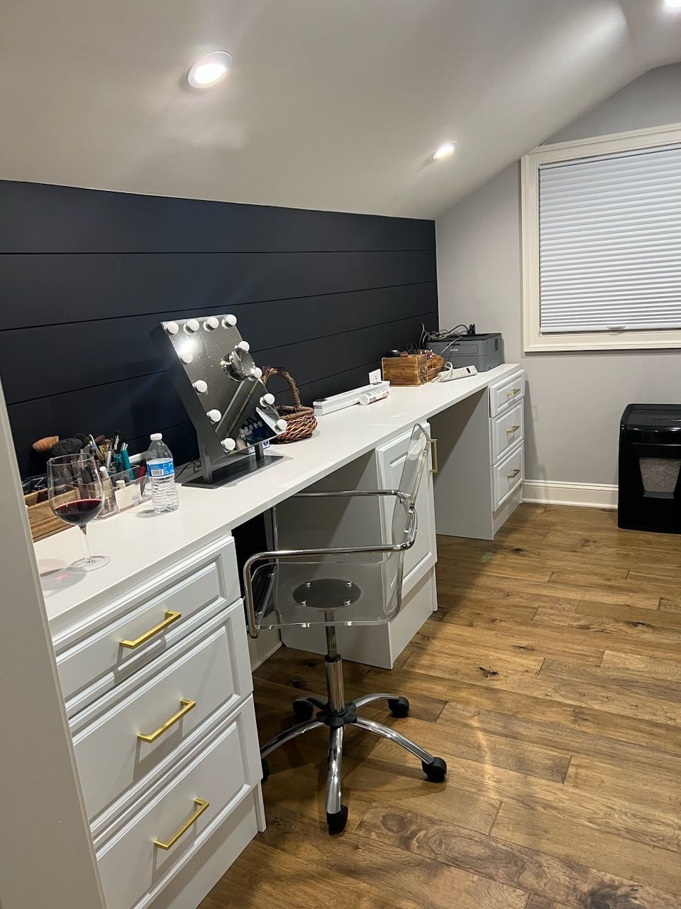 A home vanity area featuring a white desk with gold pulls, a lighted mirror, and a chair against a dark horizontal wall.