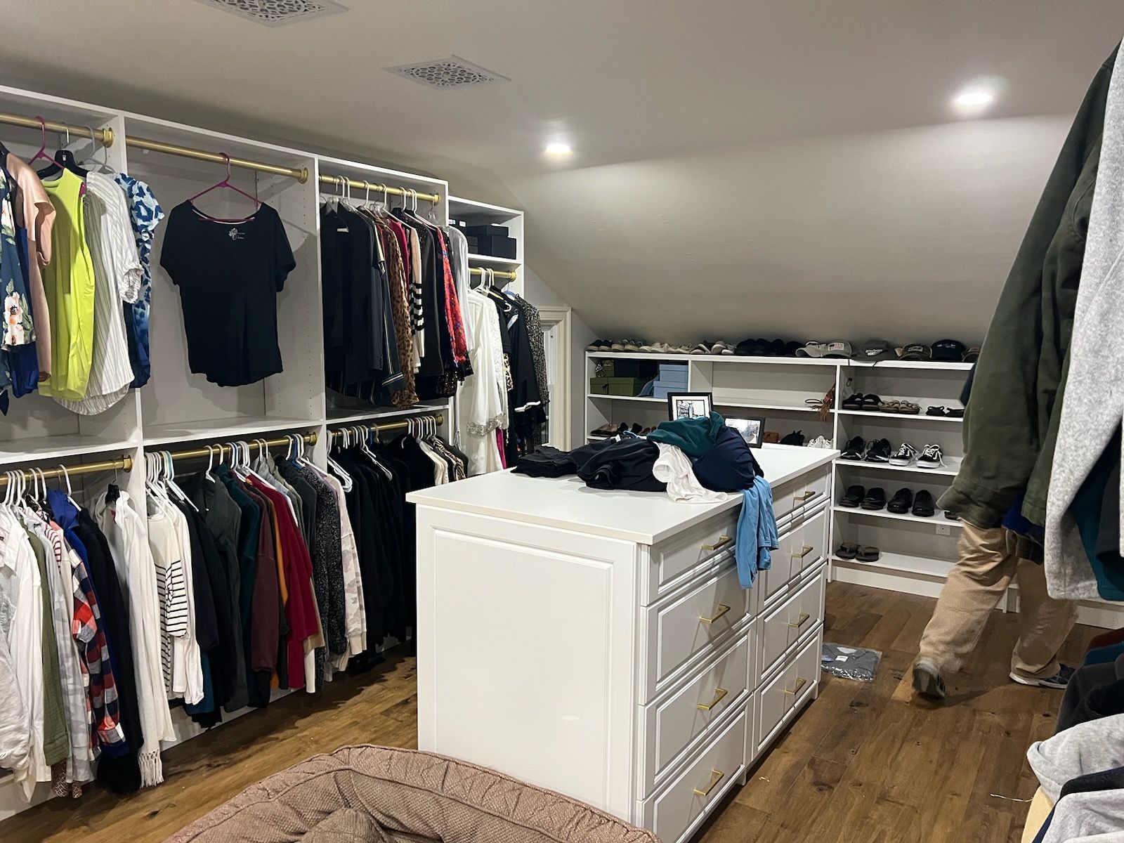 A walk-in closet with hanging clothes, a white dresser island, and built-in shelving with shoes under a sloped ceiling.
