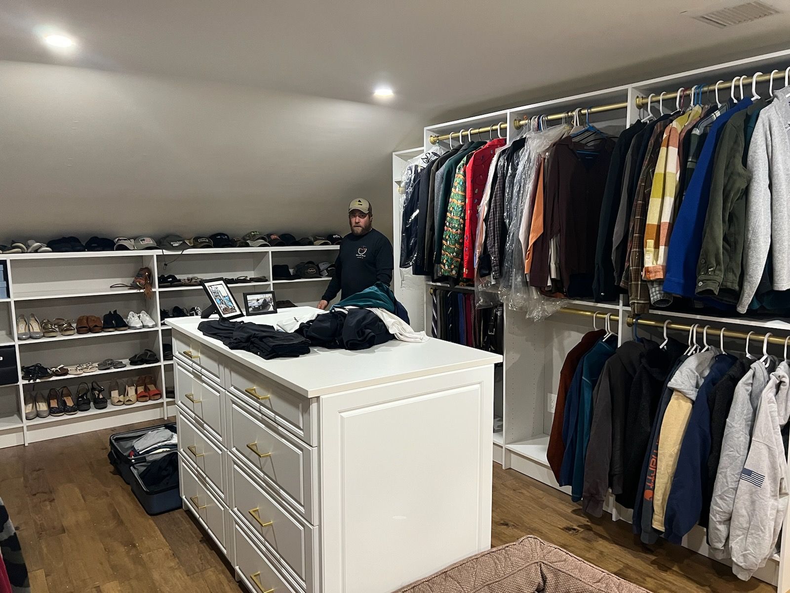 A person stands in a large walk-in closet with white shelves, hanging clothes, and a white dresser island in the center.
