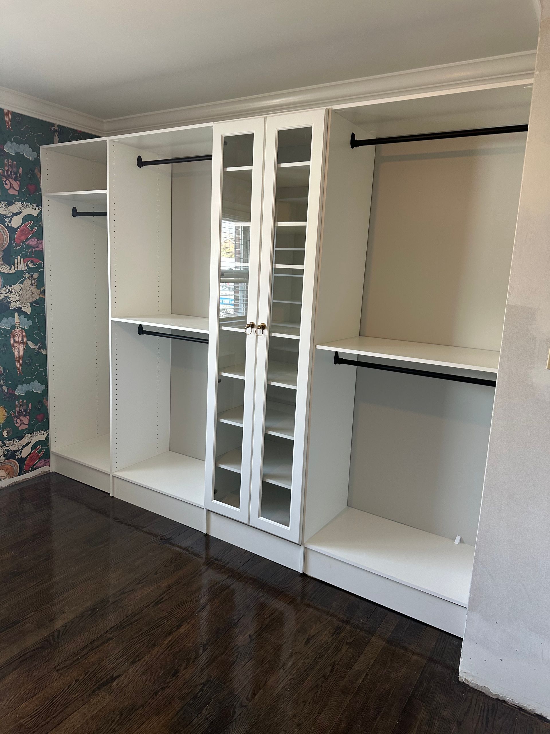 A white walk-in closet unit with shelves, hanging rods, and a central glass-door cabinet against a patterned wall.