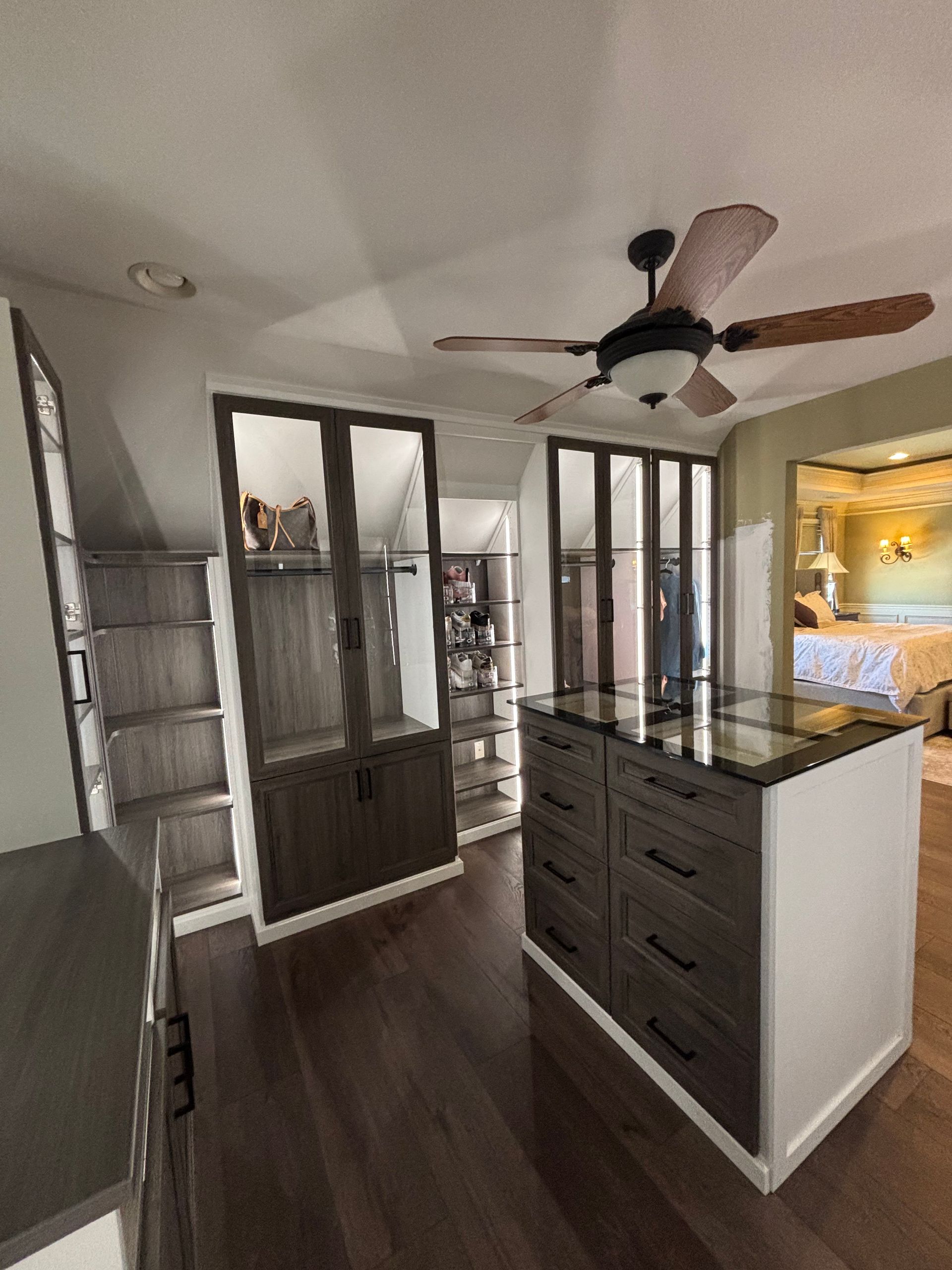 A walk-in closet featuring dark wood cabinets, glass-front wardrobe doors, an island with drawers, and a ceiling fan.