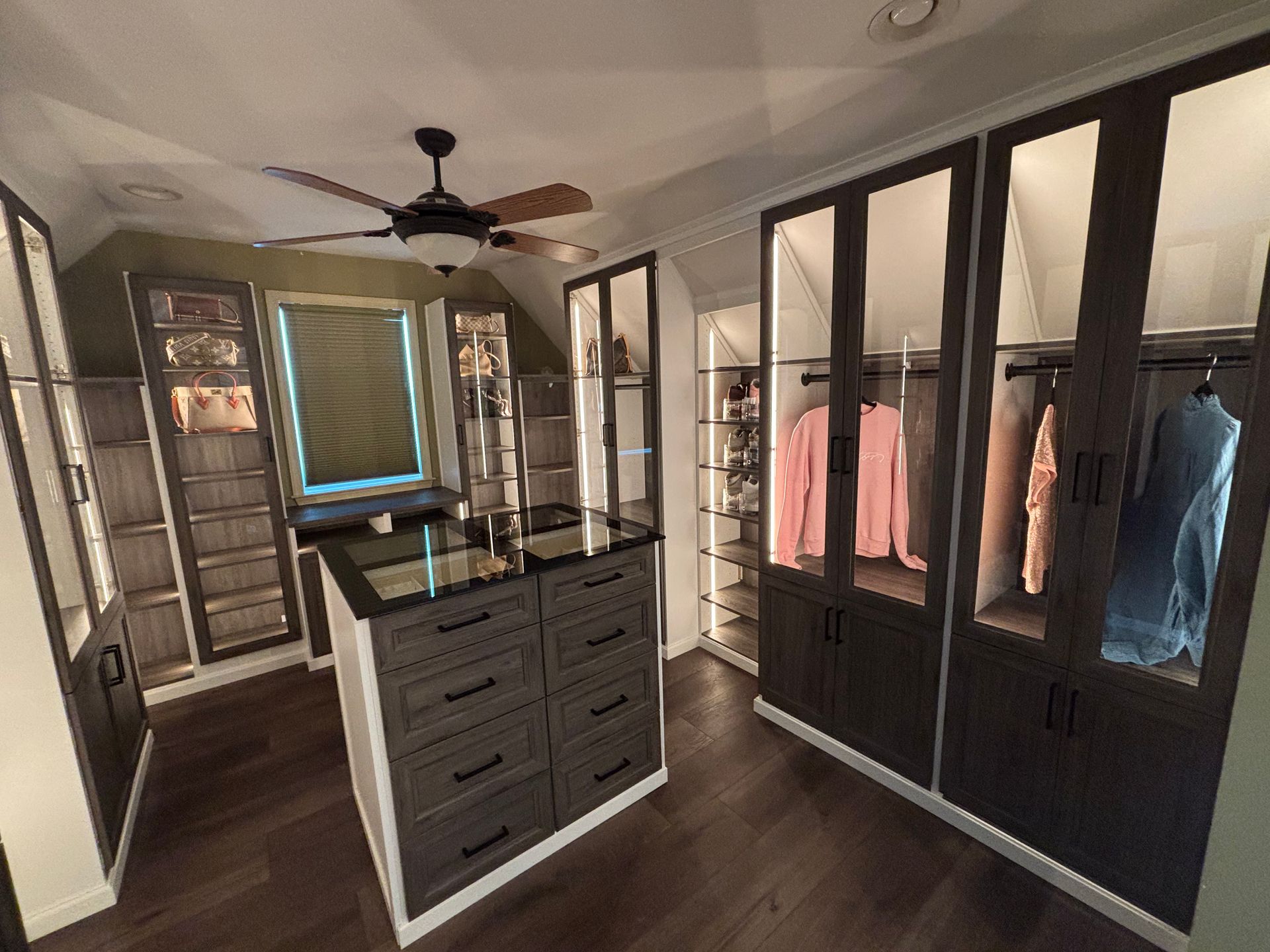 A walk-in closet featuring a dark wood center island, glass-front cabinetry with interior lighting, and dark wood floors.