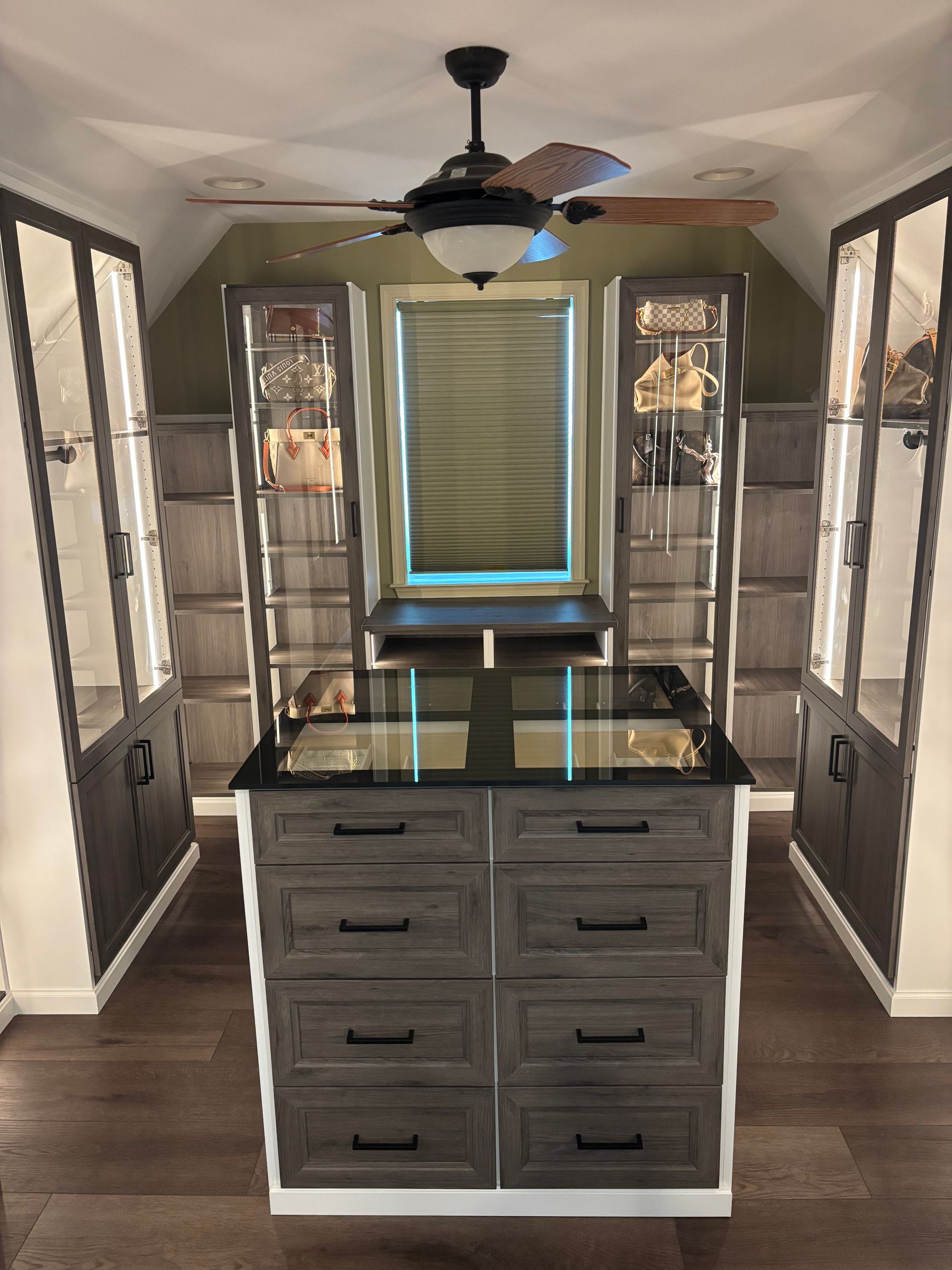 A luxury walk-in closet featuring a dark wood island, display cabinets with glass doors, and a central ceiling fan.