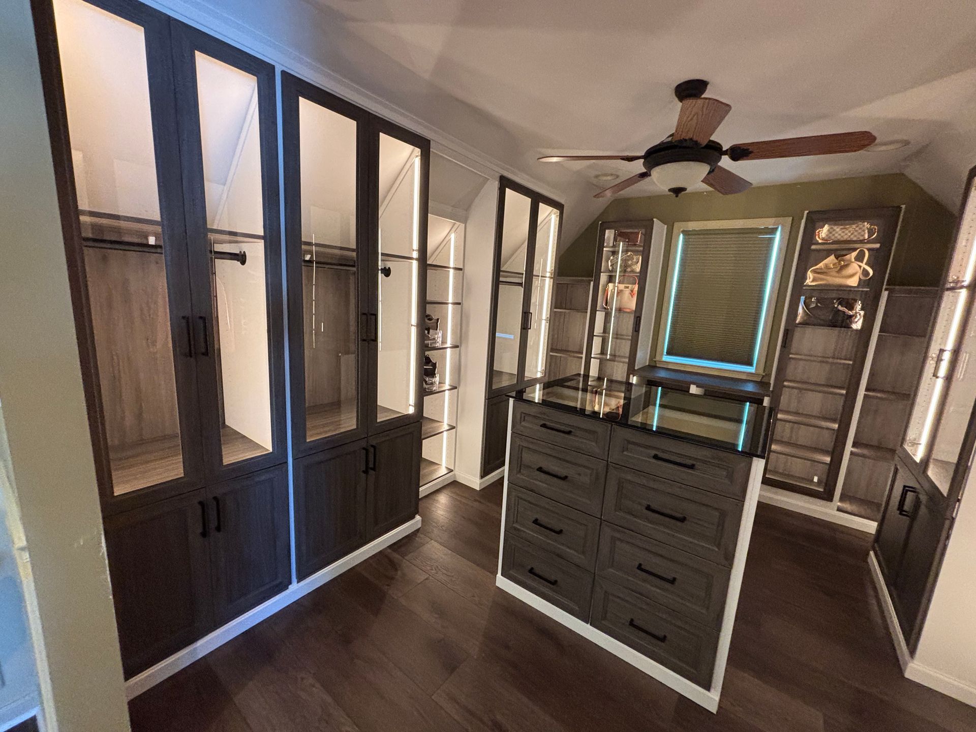 A modern walk-in closet with dark wood cabinetry, glass-fronted wardrobe doors, a central island, and a ceiling fan.
