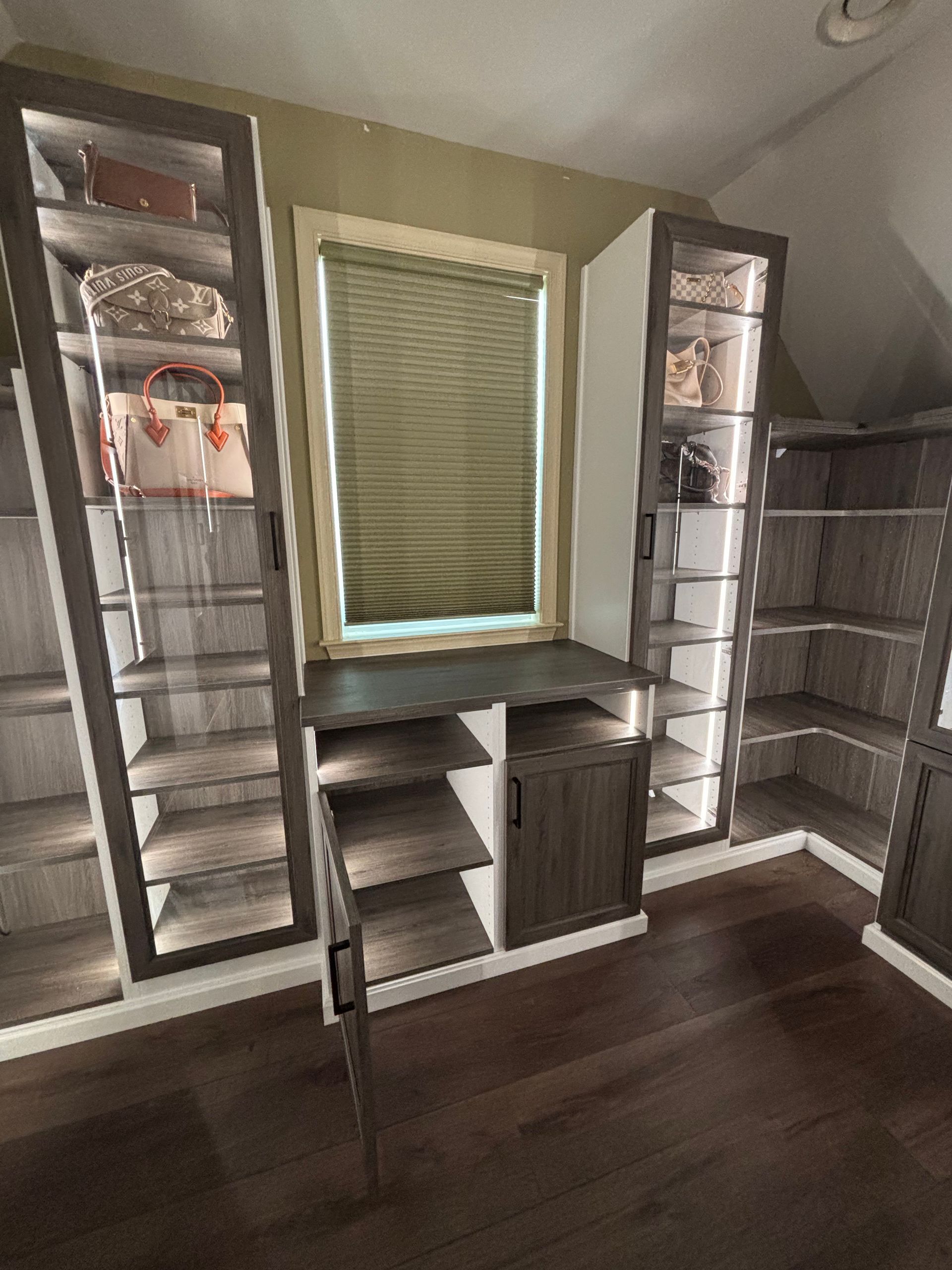 Custom wooden closet shelving and cabinetry with glass-fronted displays and internal lighting beneath a central window.