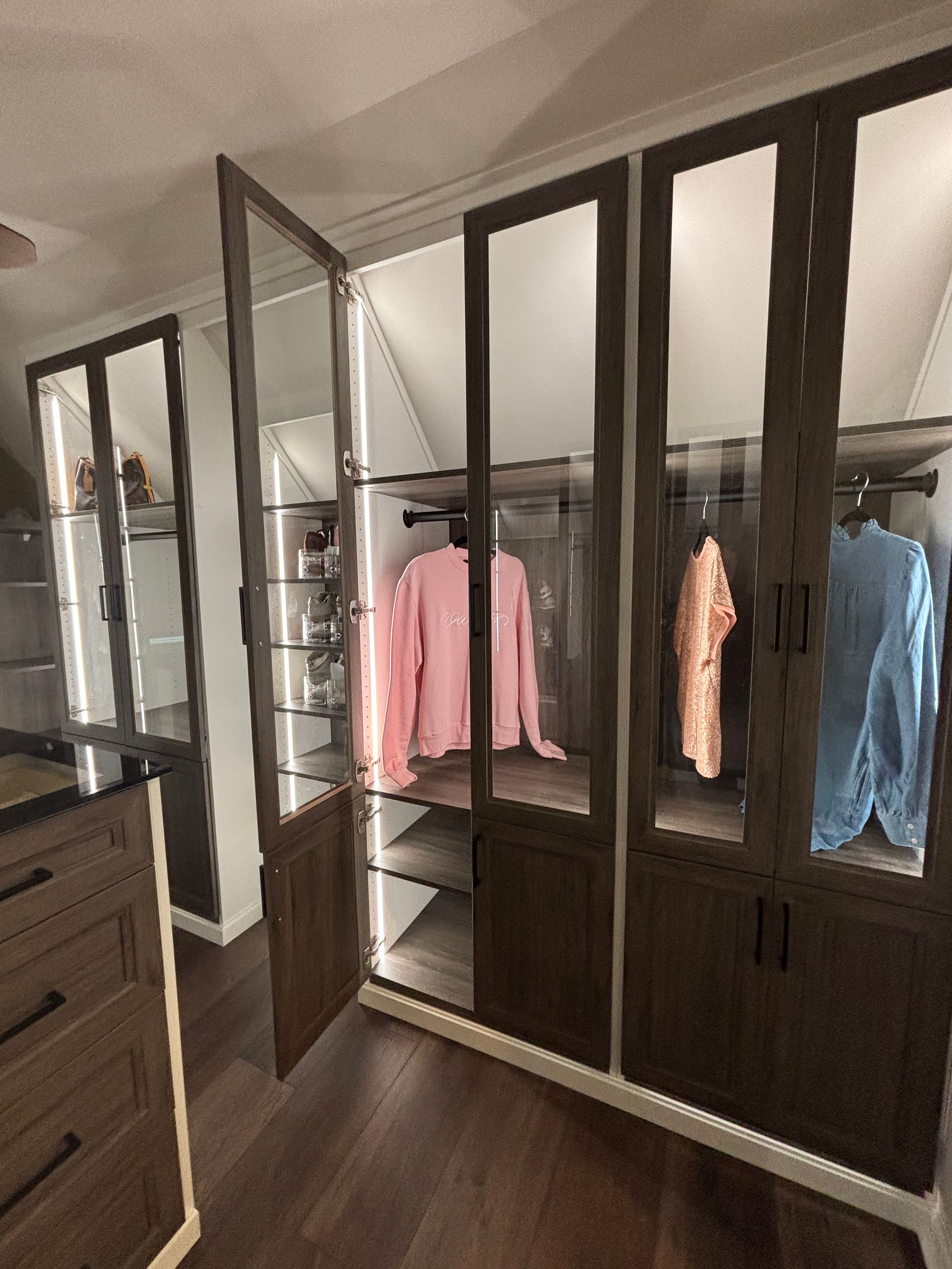A closet with dark wood frames and glass doors, featuring hanging clothing and interior lighting in a well-lit room.