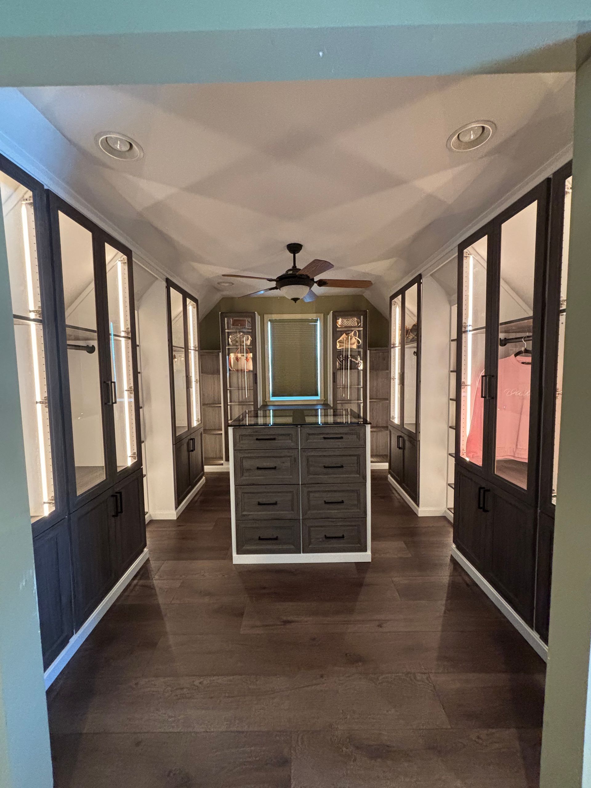 A walk-in closet with dark wood cabinetry, a central storage island with drawers, and a ceiling fan.