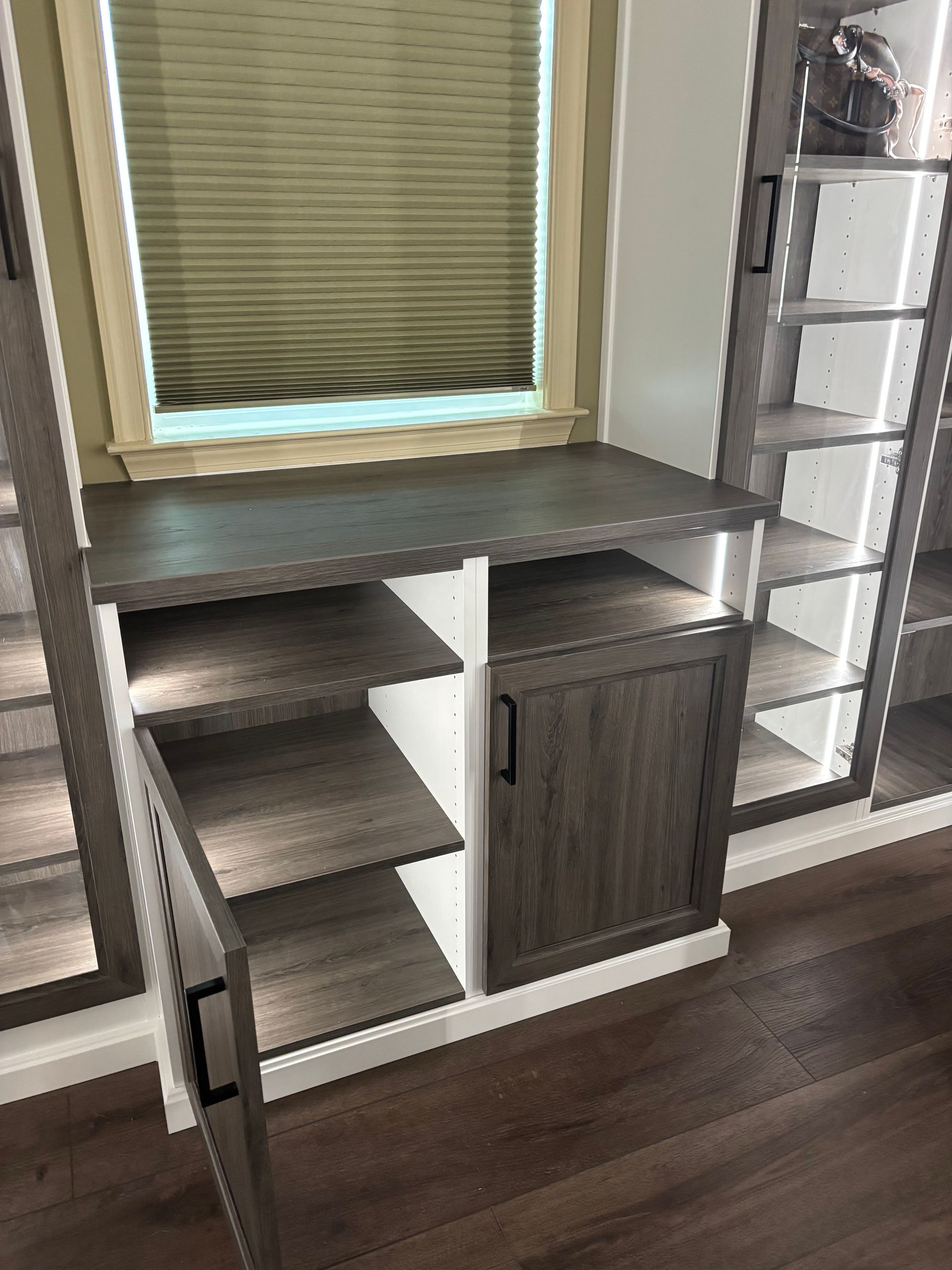 A dark wood storage cabinet with open shelving and a closed door, positioned below a window with a pleated shade.
