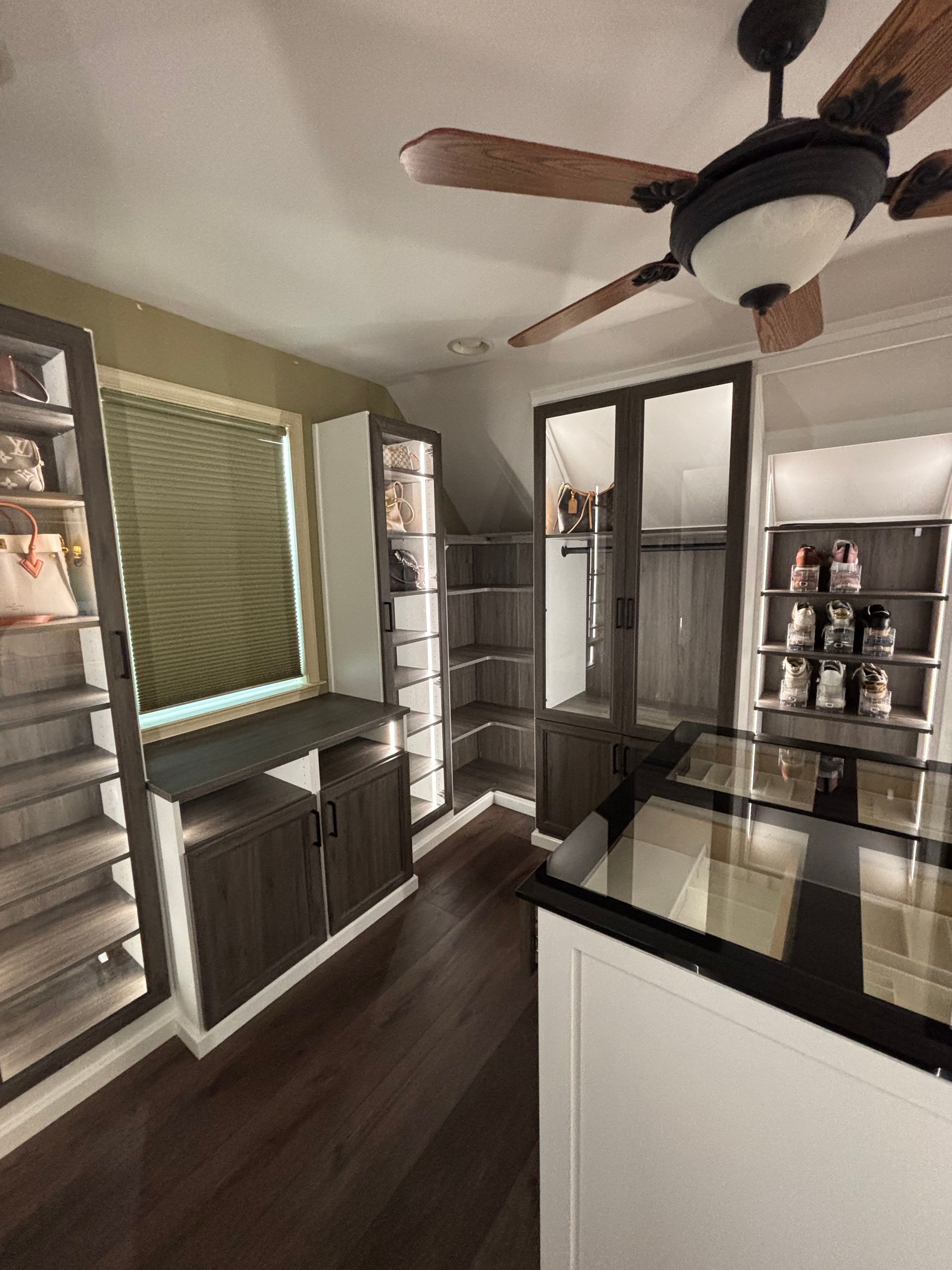 A modern, neutral-toned walk-in closet with dark wood floors, built-in shelving, glass-top center island, and ceiling fan.
