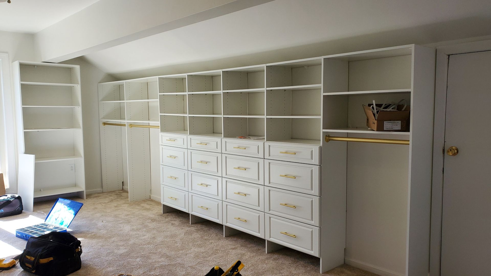 A wide, white custom closet shelving and drawer unit being installed in a room with carpet flooring.