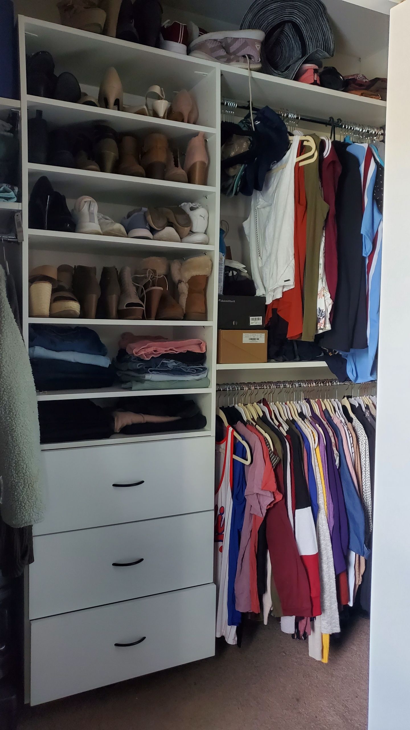 A white closet organizer with shelves filled with shoes, folded clothes, and drawers next to a rack of hanging clothing.