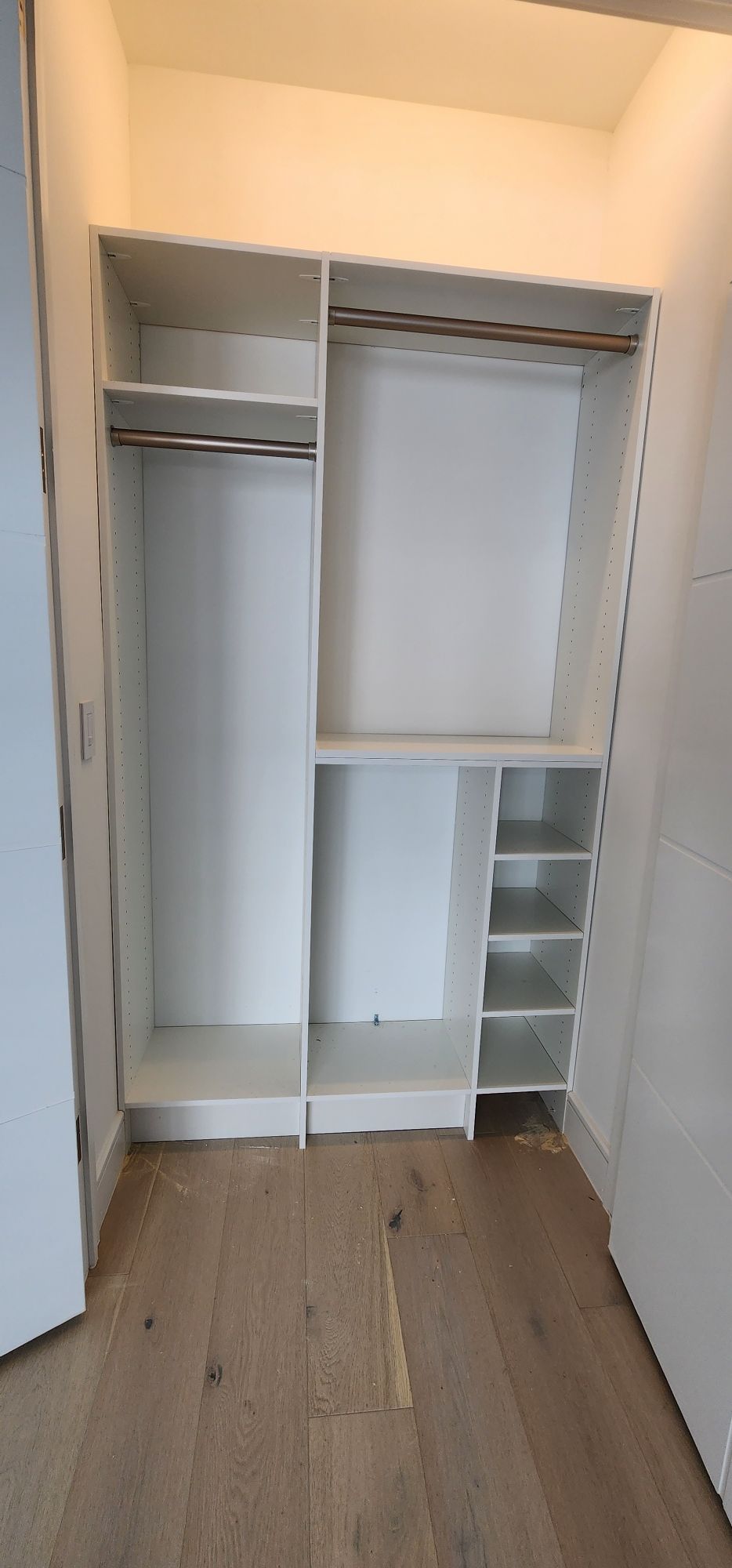 Empty white closet organizer with hanging rods and side shelving above light wood flooring.