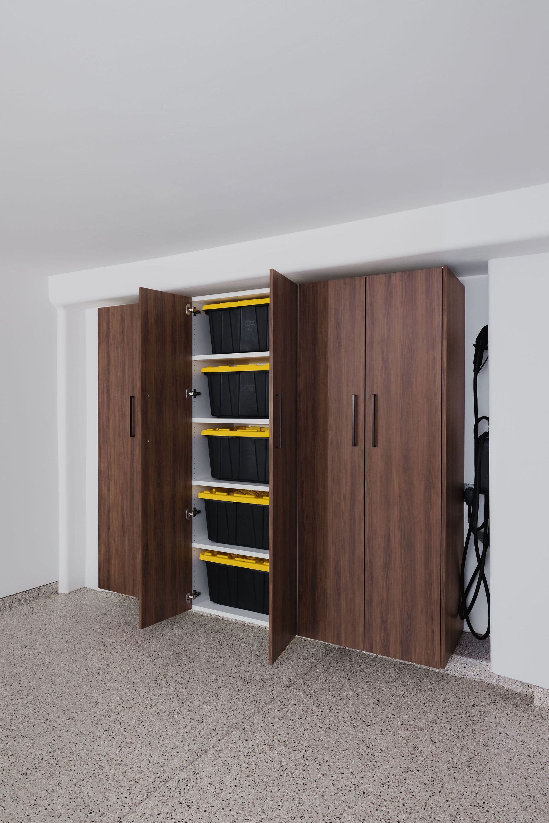 A set of dark wood floor-to-ceiling cabinets installed in a garage, one open section showing stacked storage bins inside.
