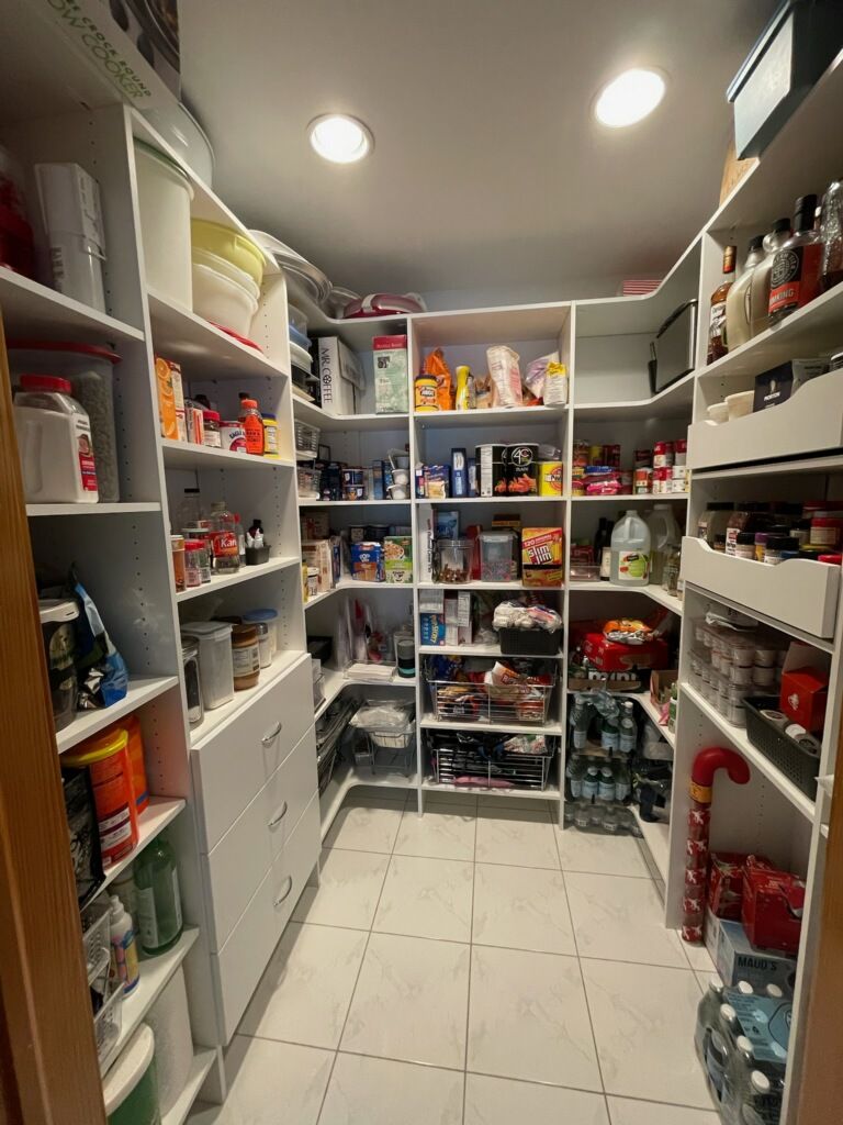 A walk-in pantry with white shelves filled with various food items, storage containers, and a tiled floor.