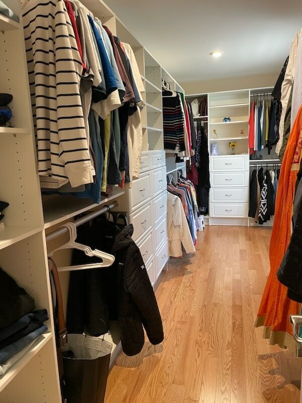 A walk-in closet with rows of hanging clothes, shelving units, and white drawers on hardwood floors.