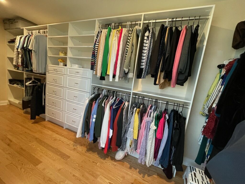 A large, white custom closet system filled with hung clothes and drawers, situated in a room with hardwood floors.