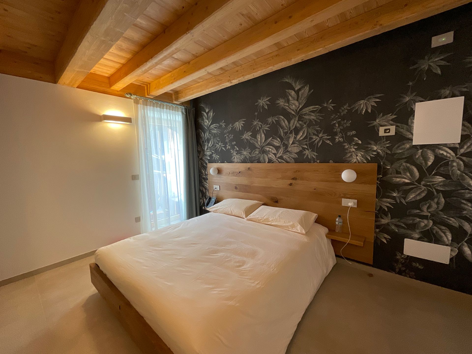 Camera da letto con struttura del letto in legno, carta da parati floreale e travi in ​​legno a vista sul soffitto.