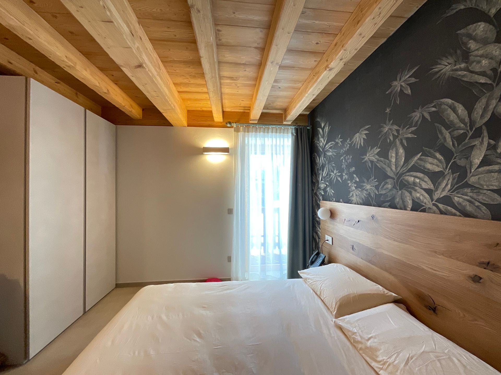 Camera da letto con travi in ​​legno, letto bianco, carta da parati floreale scura e armadio a muro.