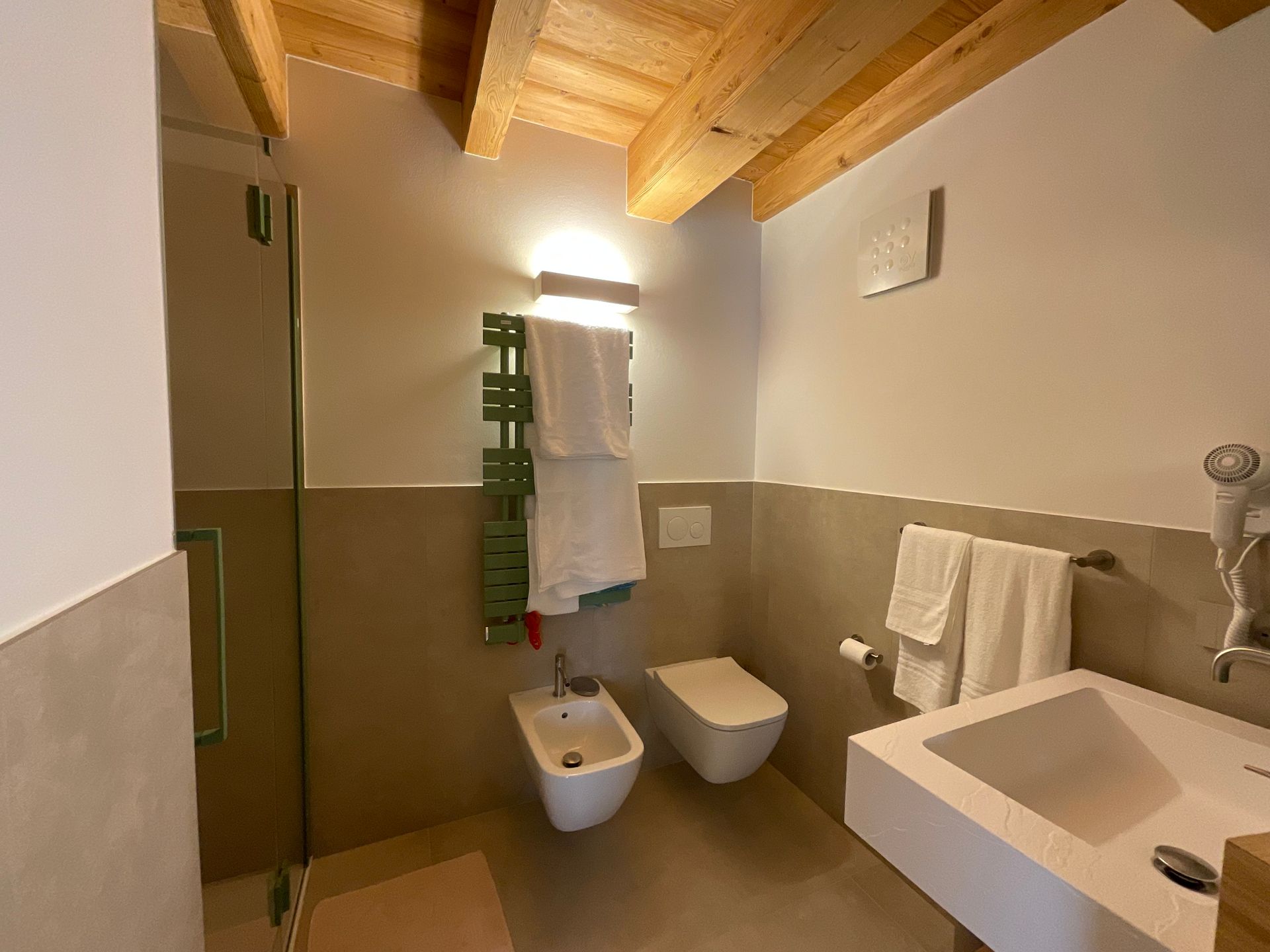 Bagno con sanitari bianchi, doccia e portasciugamani verde. Soffitto in legno e pareti dai toni neutri.