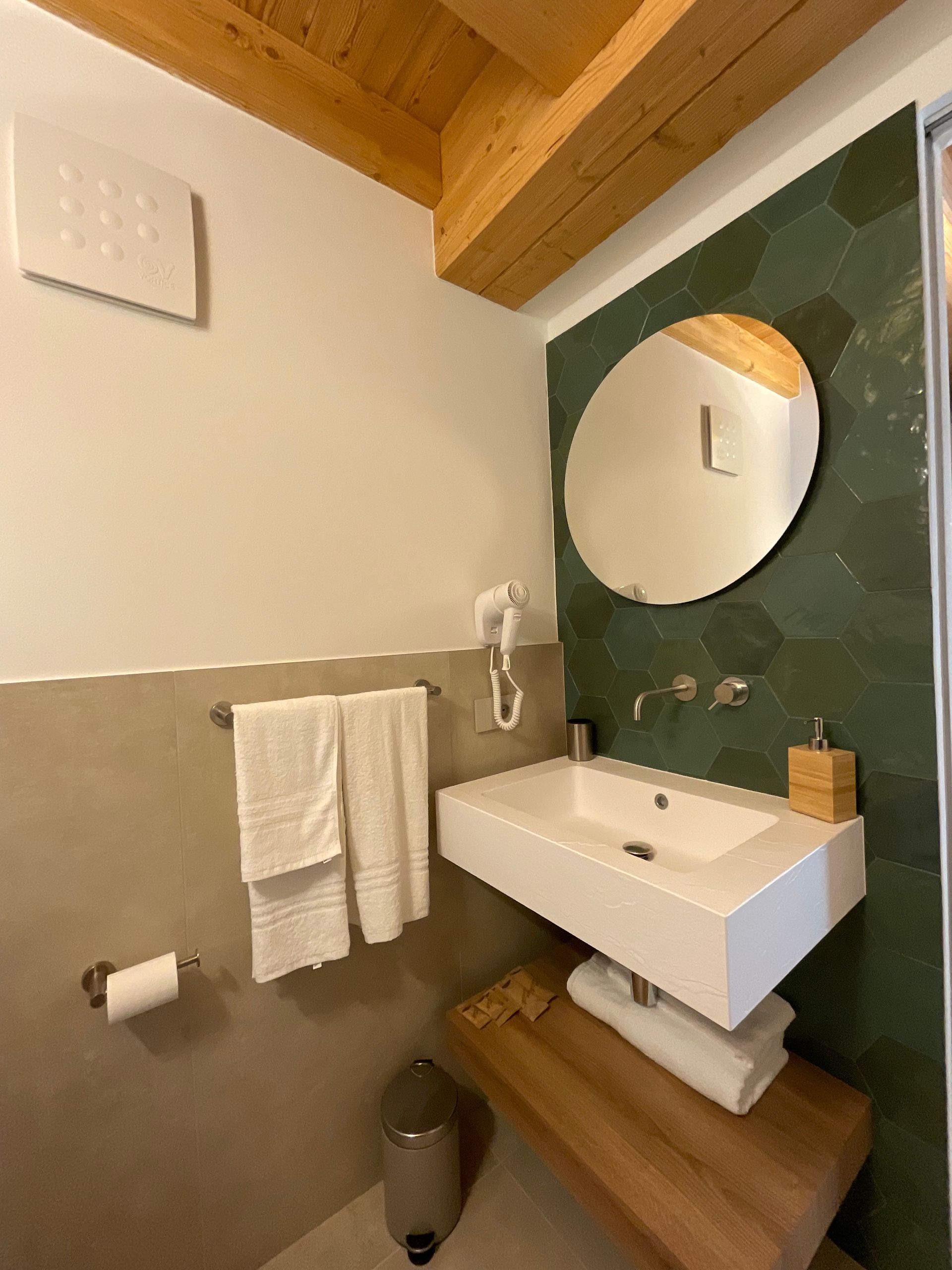 Bagno con lavandino bianco, specchio rotondo, parete piastrellata verde, asciugamani e dettagli in legno.