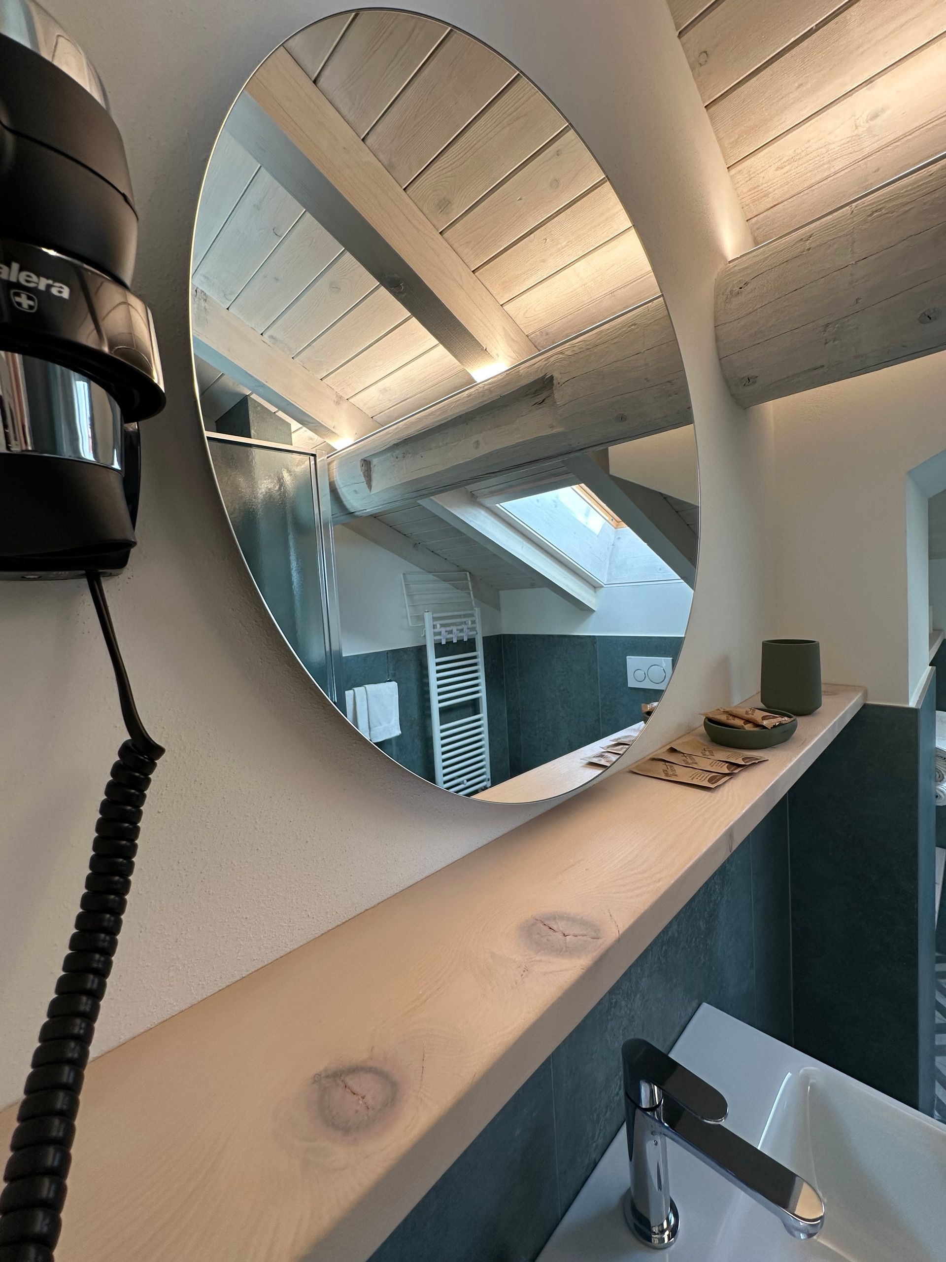 Specchio ovale che riflette un bagno con lavandino, mensola e travi a vista. Un asciugacapelli è appeso alla parete.