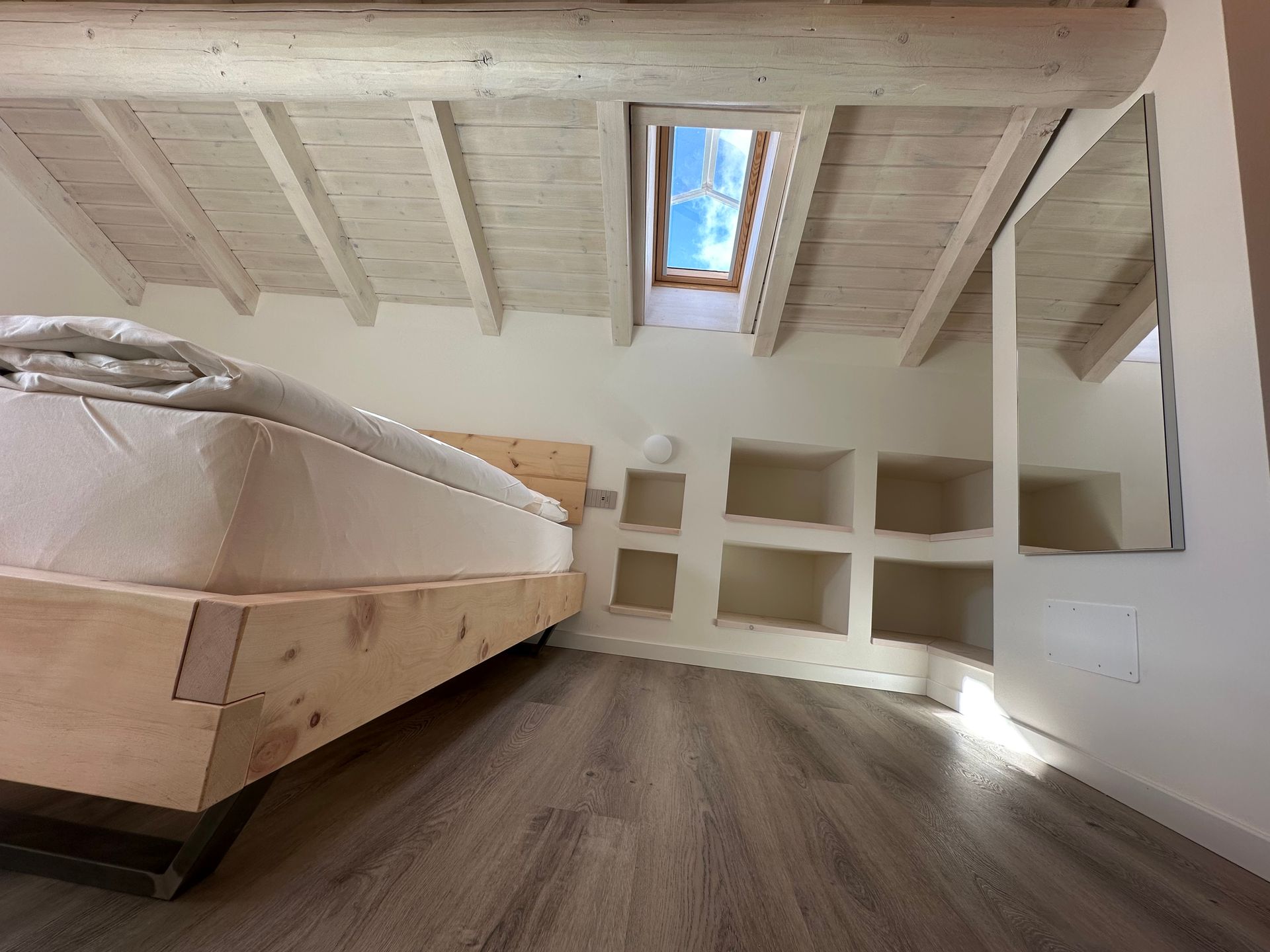 Camera da letto con letto in legno, lucernario e scaffali integrati sotto un soffitto con travi bianche.