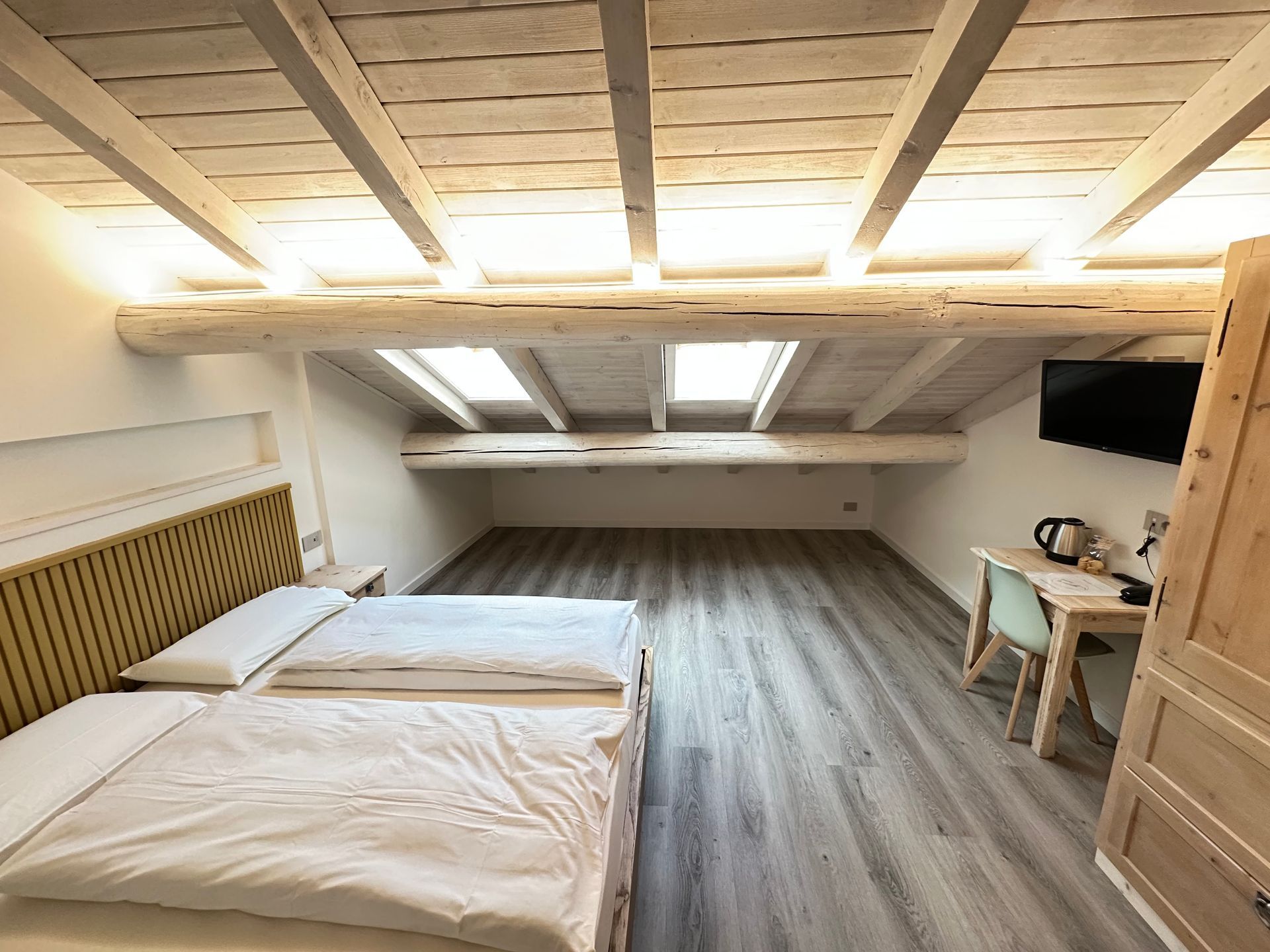 Camera da letto con due letti, travi in ​​legno, lucernari, scrivania e TV. Legno chiaro e pareti bianche.