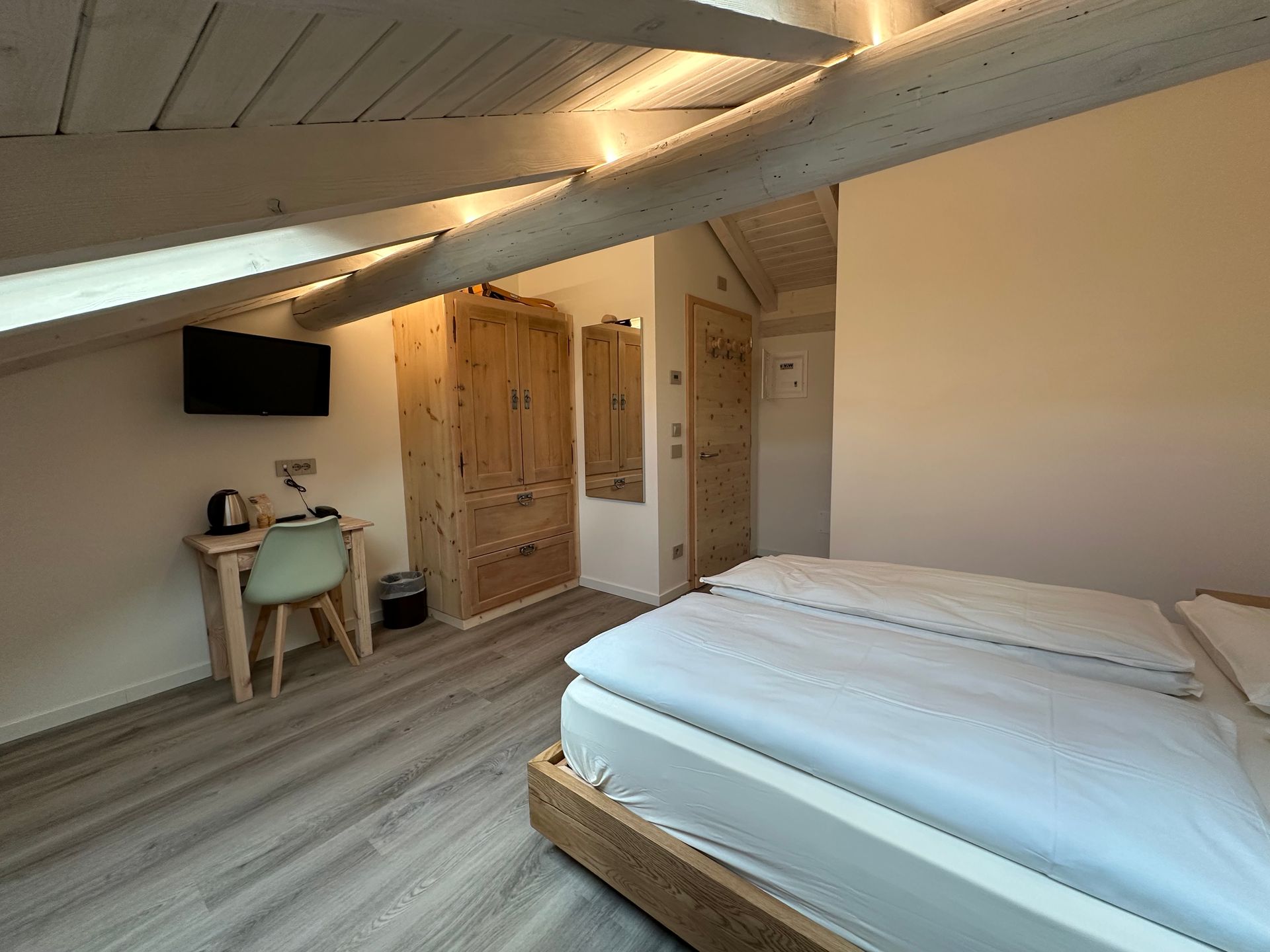 Camera da letto con soffitto spiovente, letto, scrivania, armadio e porta. Travi in ​​legno e pavimento in legno chiaro.