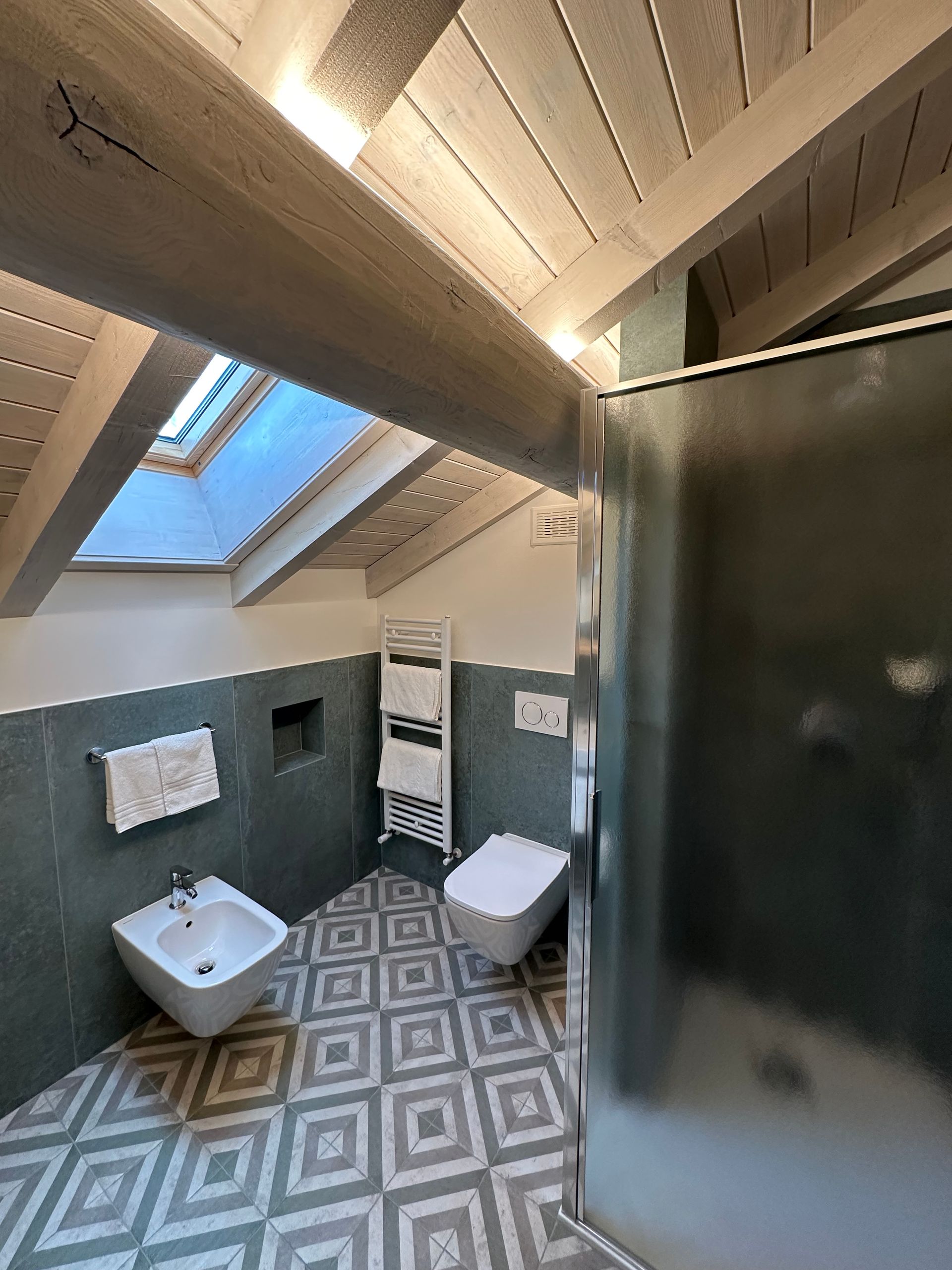Bagno con lucernario, travi a vista, pareti piastrellate color verde acqua, pavimento geometrico, bidet, WC e doccia in vetro smerigliato.