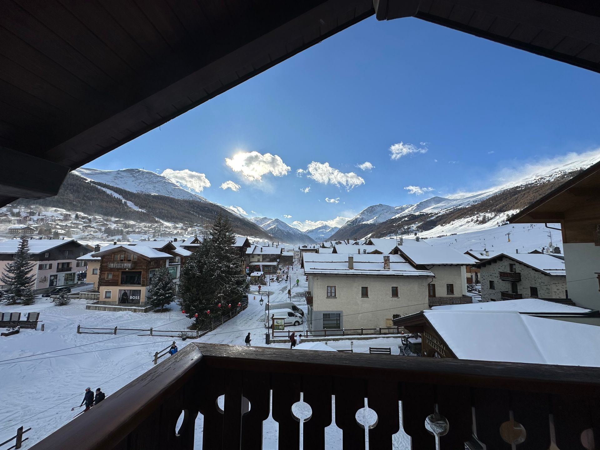 Vista di un villaggio di montagna innevato da un balcone, con edifici, una piazza innevata e cielo azzurro.